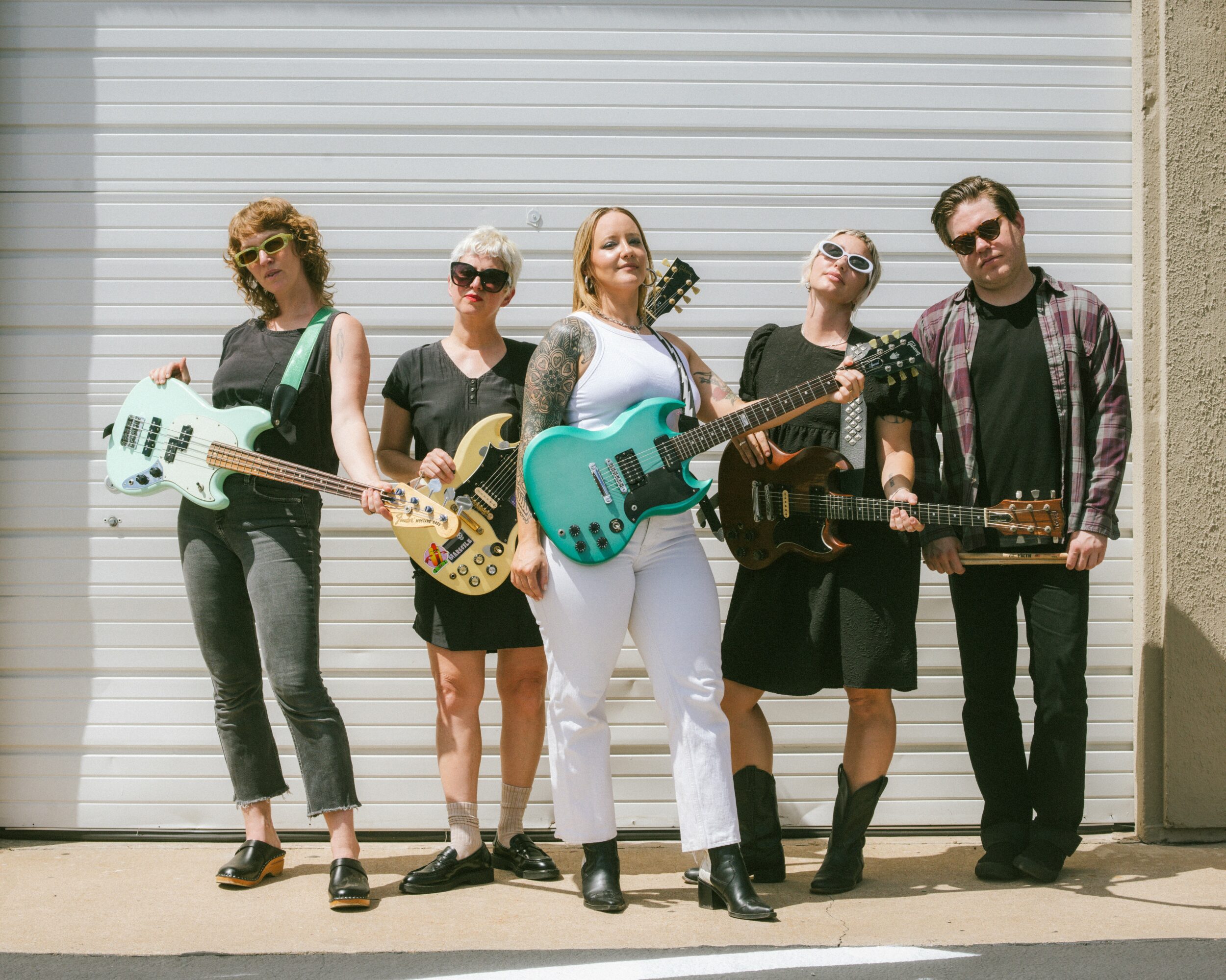 Group of five women standing outdoors with musical instruments, in front of a metal garage door, smiling and posing.