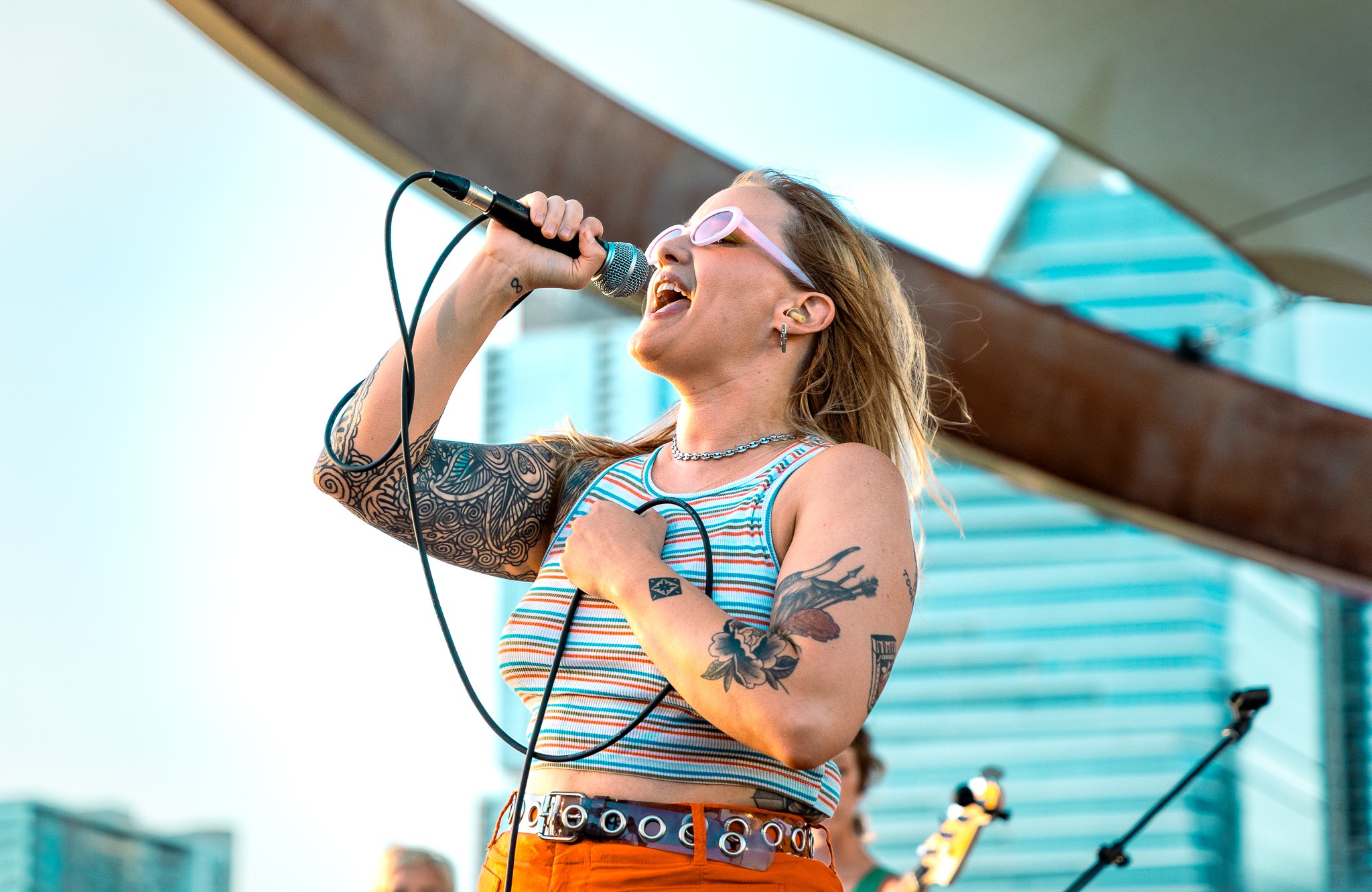 Woman singing into a microphone outdoors with modern buildings in background, wearing sunglasses and a striped top.