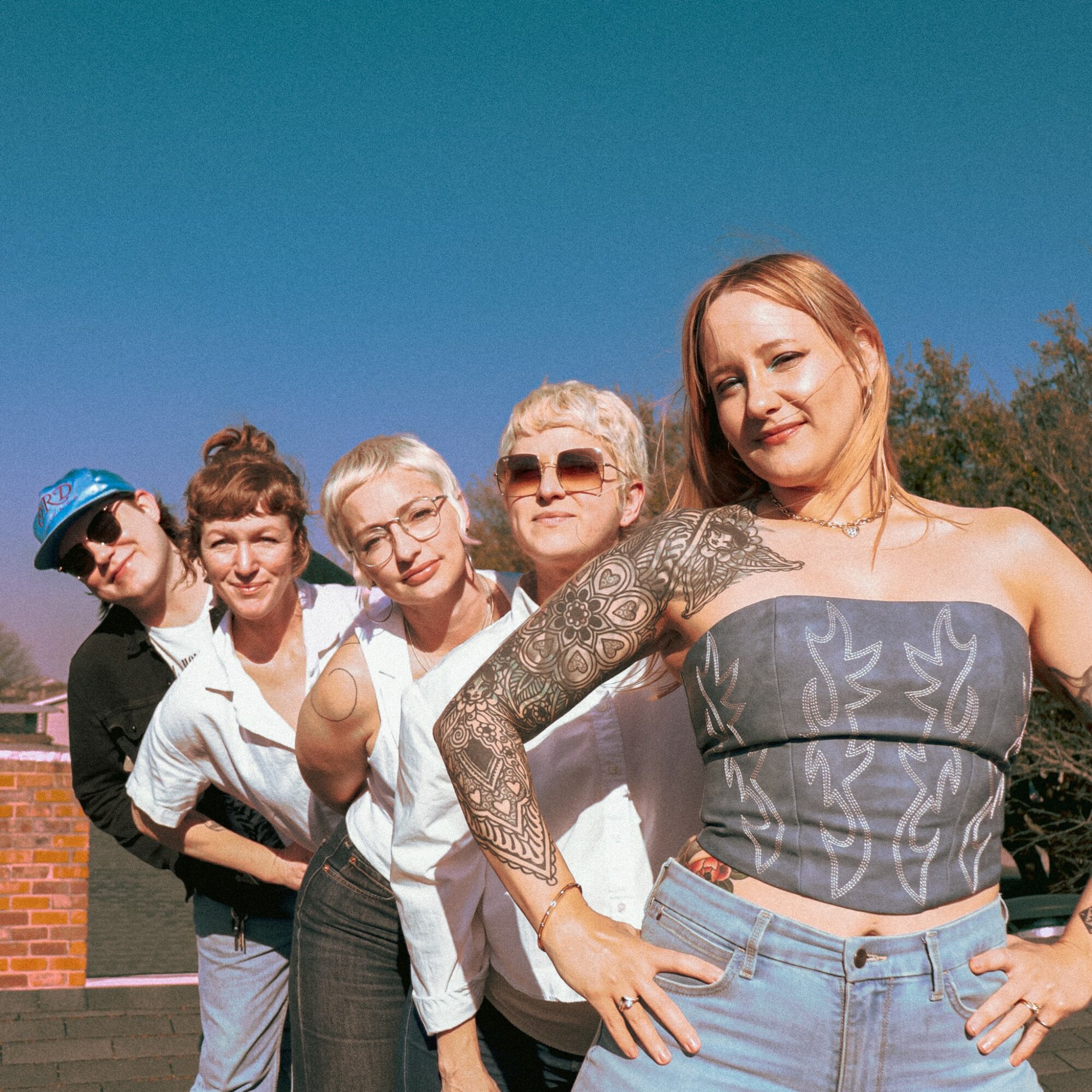 Group of five women outdoors, smiling, with trees and a building in the background.