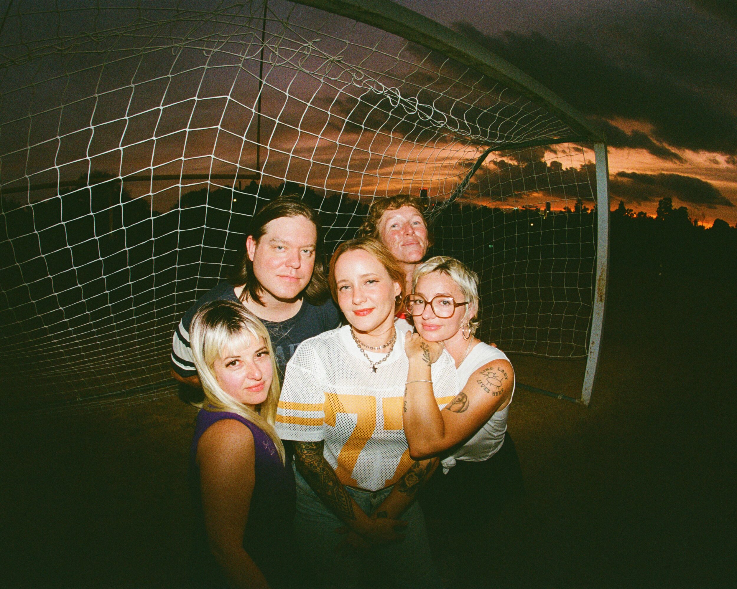 Group of five women standing together in front of a soccer goal at sunset, smiling and embracing each other.