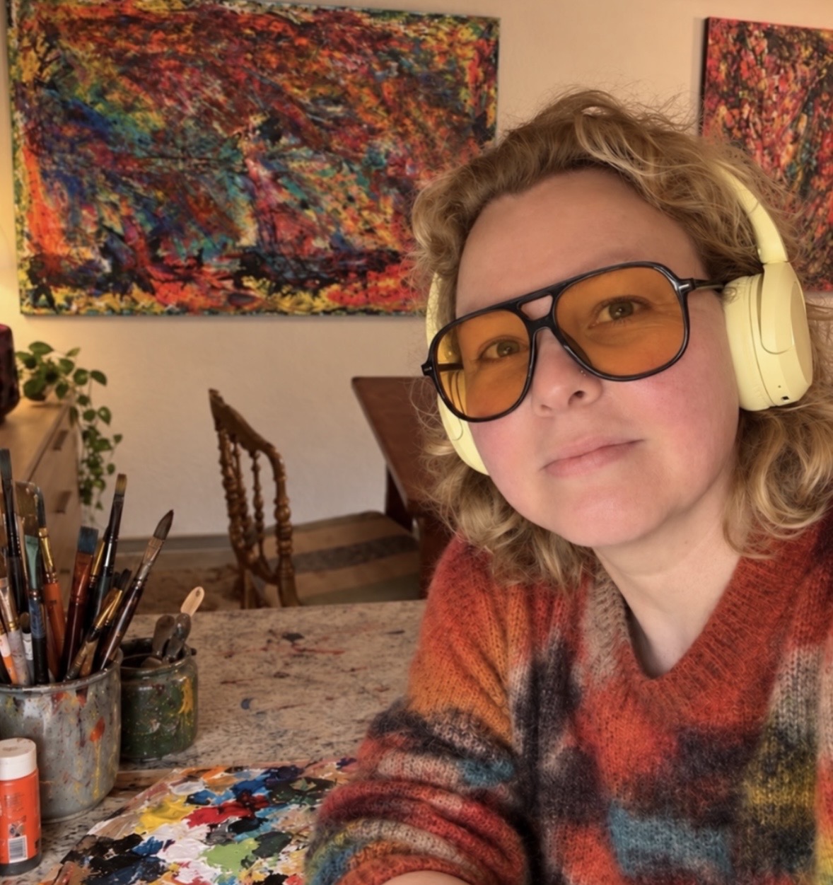 Person with headphones and sunglasses in an art studio with colorful paintings and art supplies.