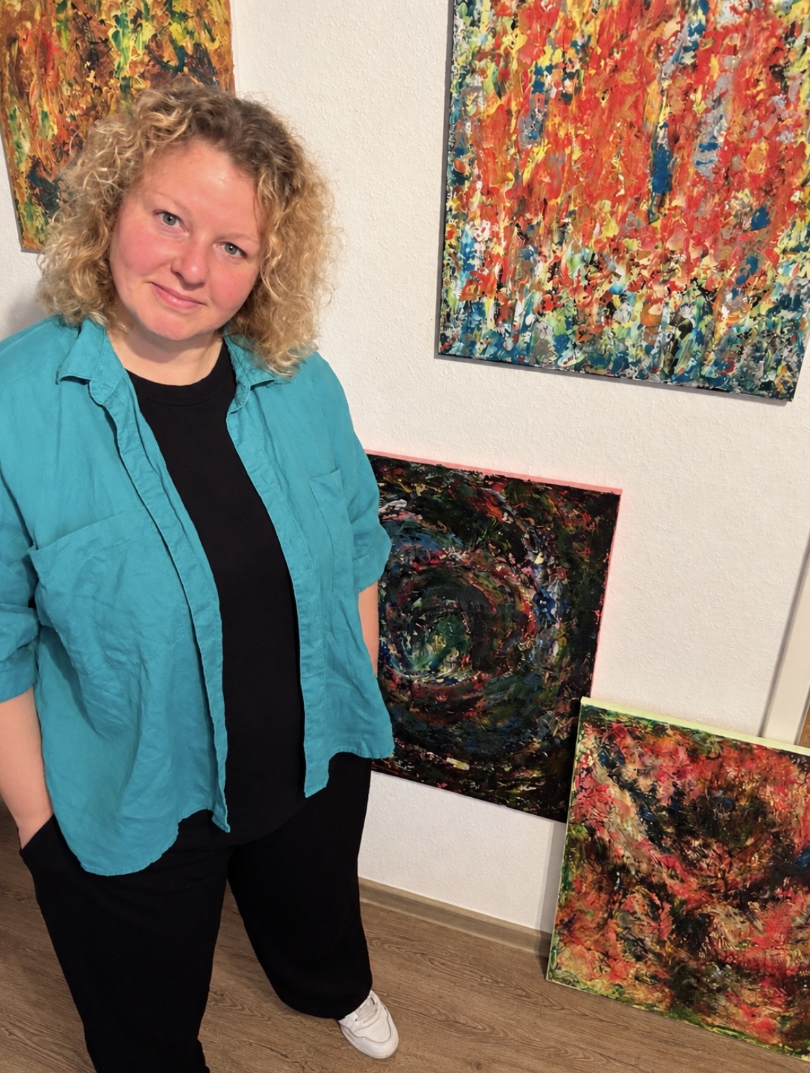 Woman with curly hair in a teal jacket standing near colorful abstract paintings on a white wall, wooden floor visible.