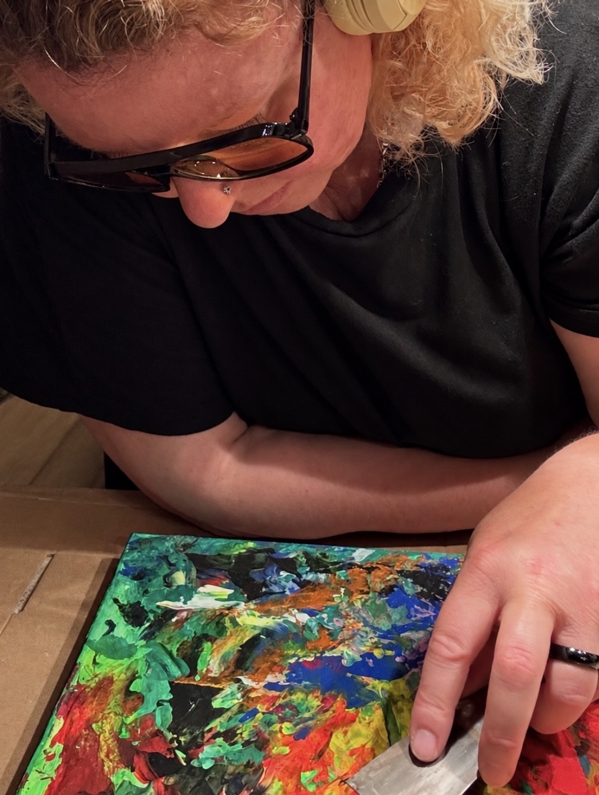 Person with curly hair and glasses looking at a colorful abstract painting on a table.