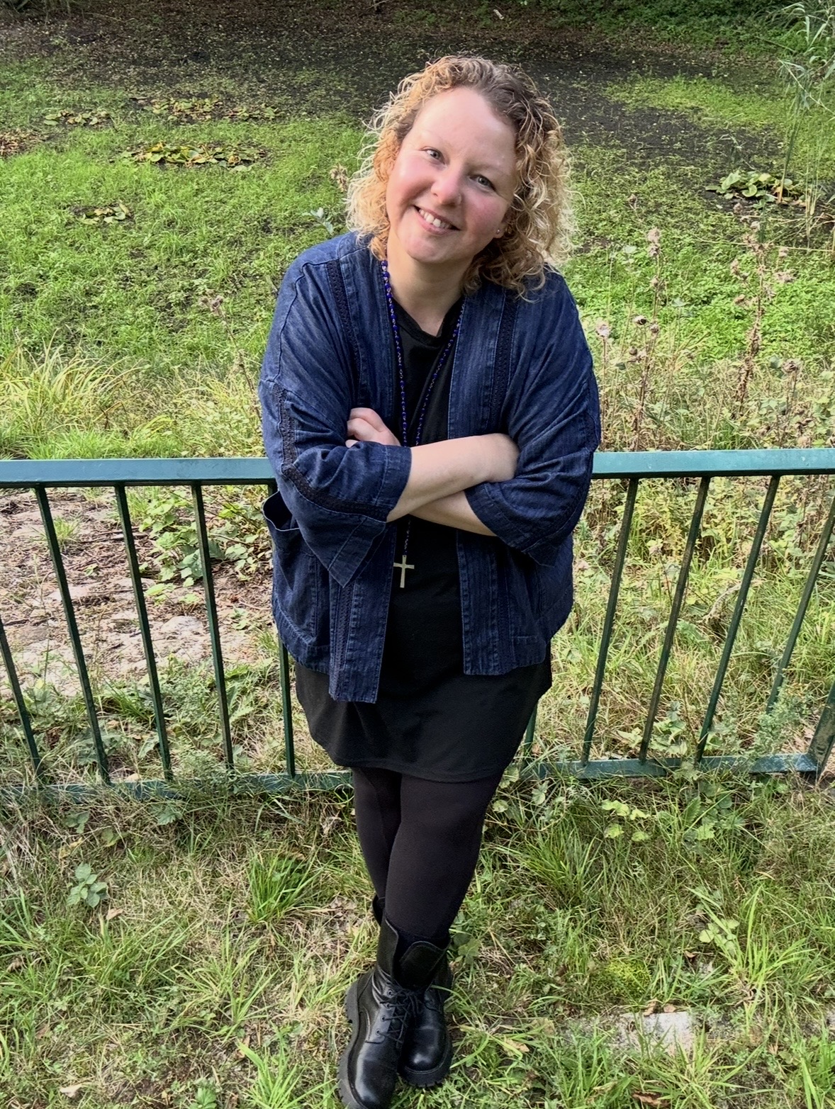 Woman with curly hair smiling, arms crossed, standing outdoors in front of a metal fence, wearing a dark dress, jacket, and boots.