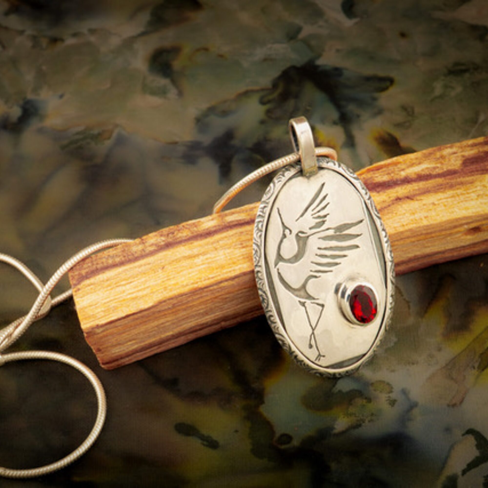 Oval pendant with engraved bird and a red gemstone, hanging on a string, resting on a wooden stick over a water surface.