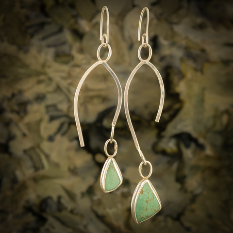 Pair of dangling earrings with curved metal hooks and green stone pendants, against a blurred background.