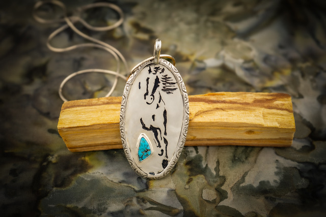 Oval pendant with abstract black lines and a blue accent, hanging on a string, placed on a wooden piece, with a textured background.