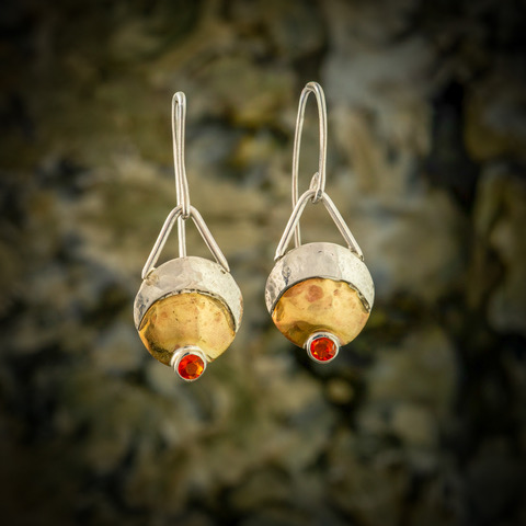 Silver earrings with gold and red gemstone accents hang against a blurred natural background.