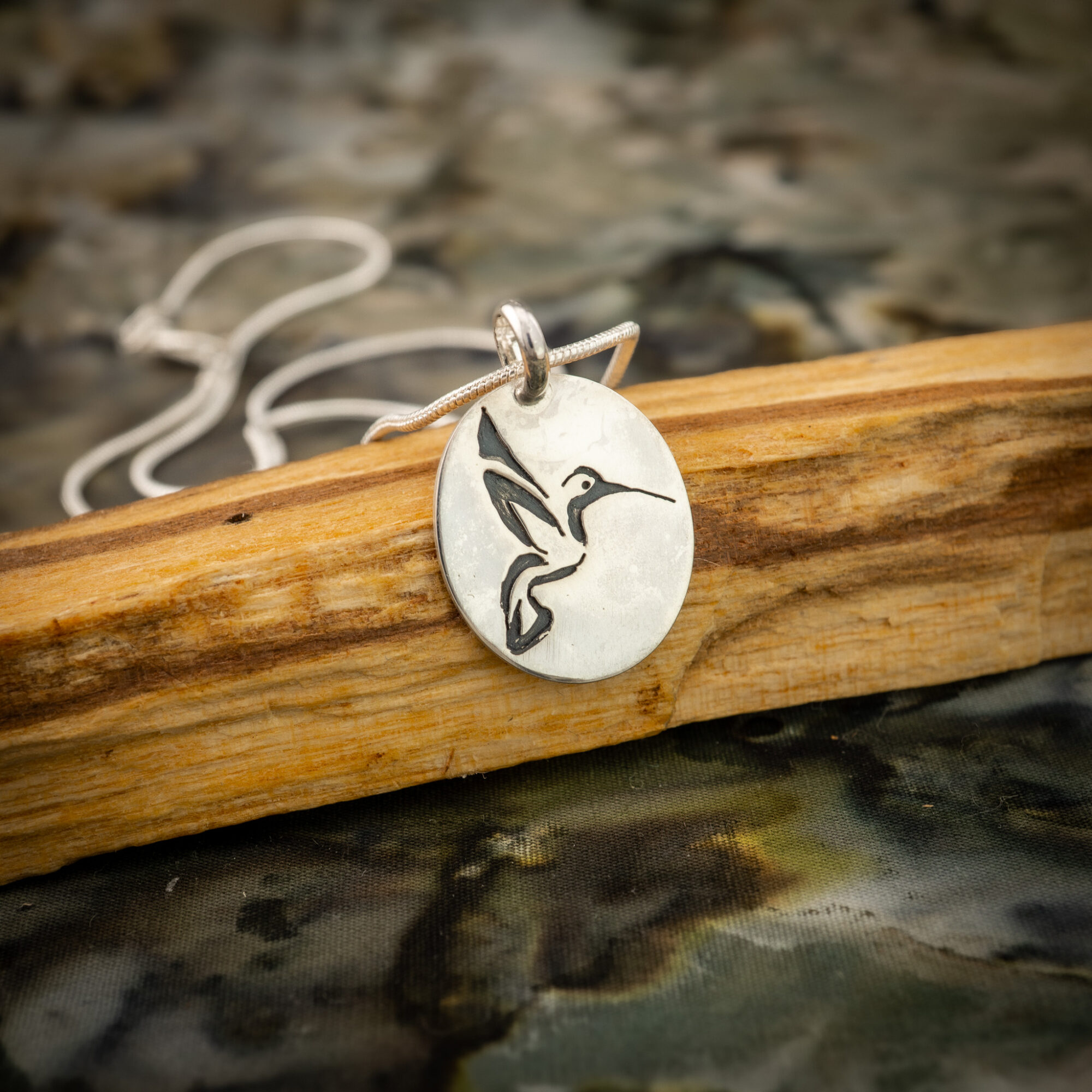 Silver pendant with a bird design, hanging on a string, resting on a wooden surface with water below.