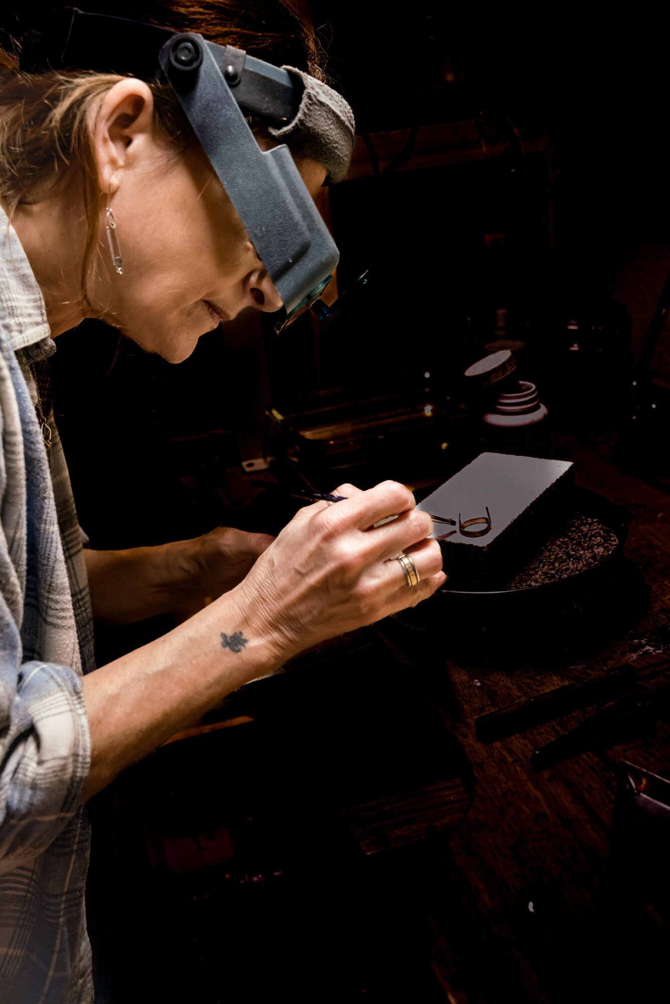 Person wearing magnifying glasses examines an object with a tool, in a dark workspace.
