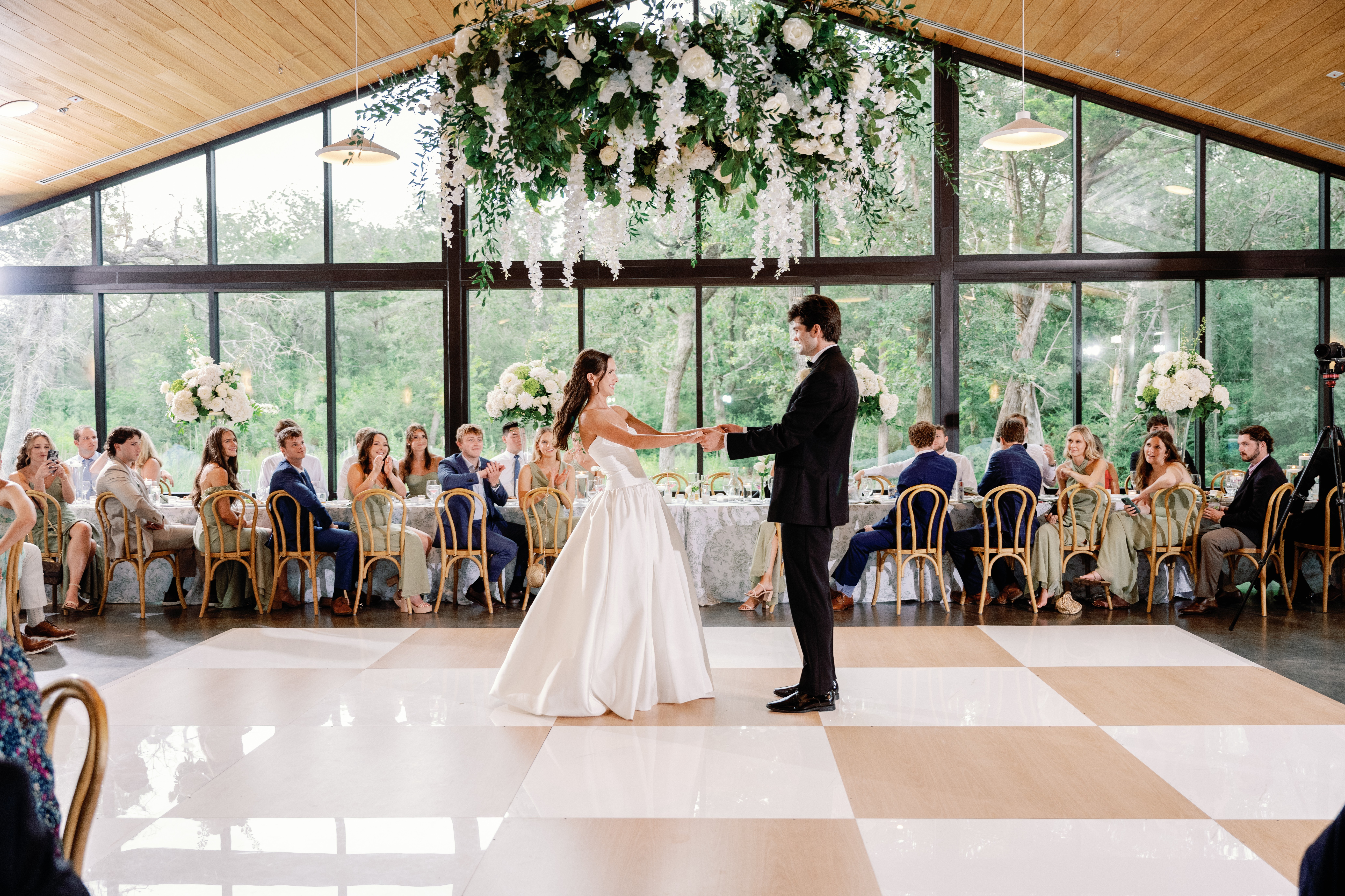 Bride and groom dance in a decorated venue with guests seated at tables, large windows, and hanging floral arrangements.