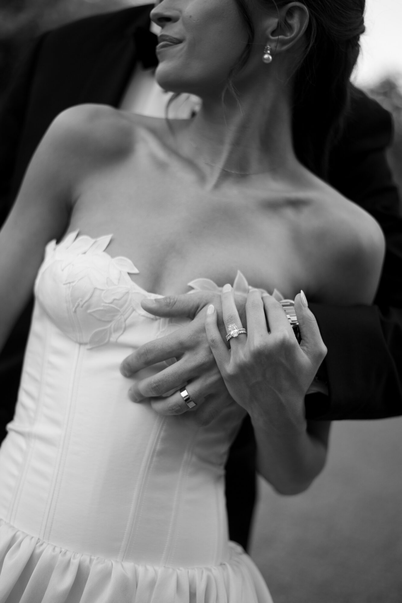 Woman in a strapless dress with jewelry, holding hands, with a person behind her, in black and white.