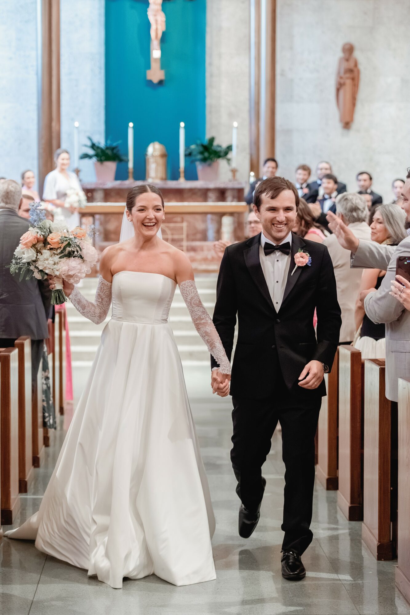 Bride and groom walking down the aisle holding hands in a church, smiling, with guests seated on both sides.