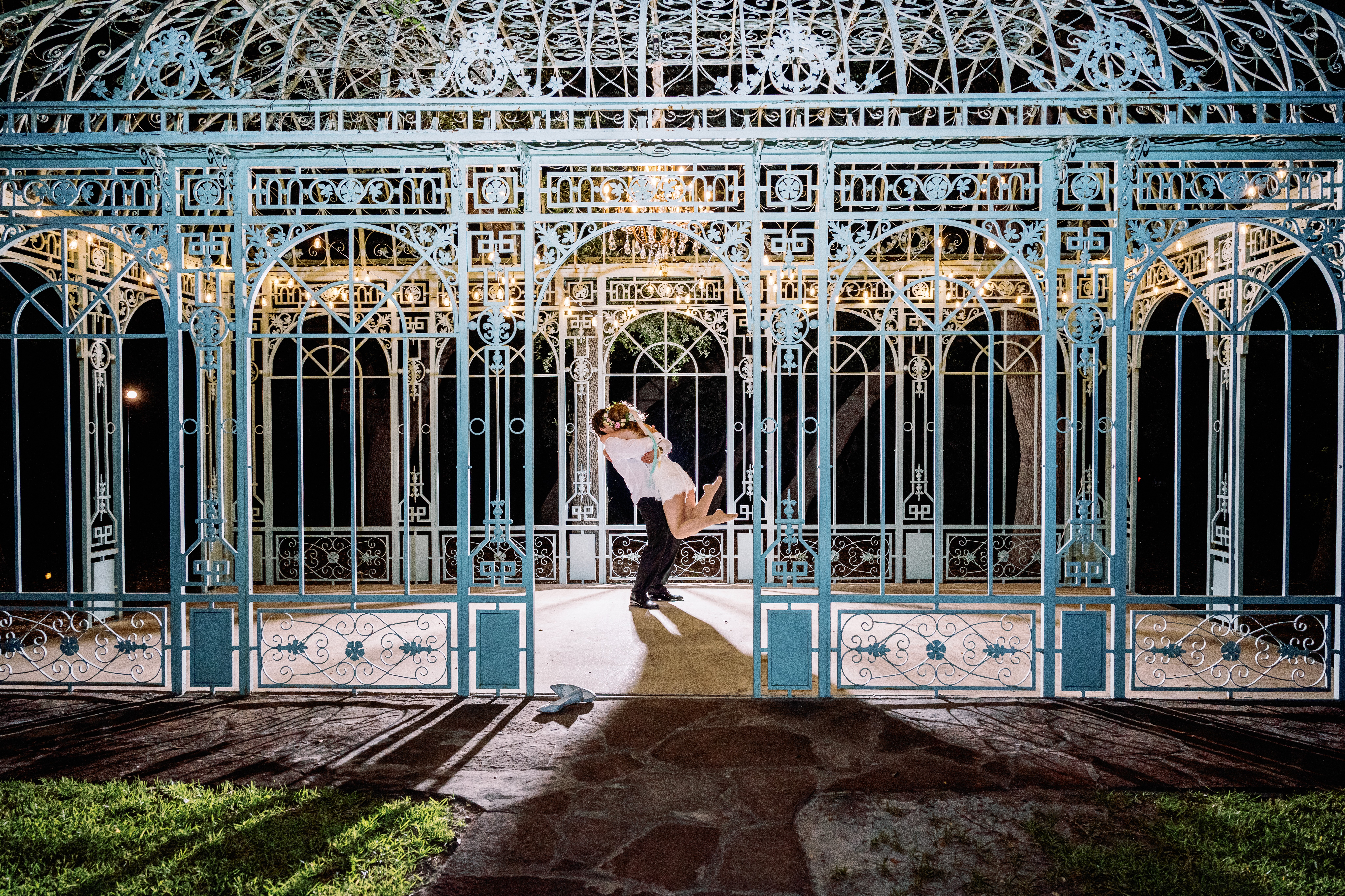 Person dancing inside a decorative, blue metal gazebo at night, with grass and stone pathway in foreground.