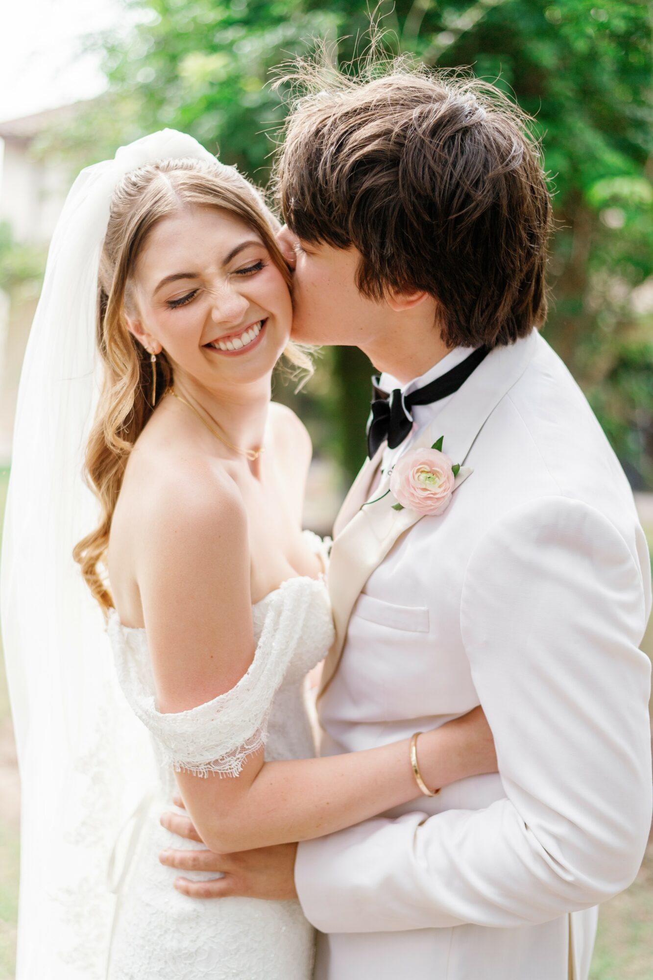 A smiling bride and groom share a kiss outdoors, with greenery in the background, during their wedding celebration.