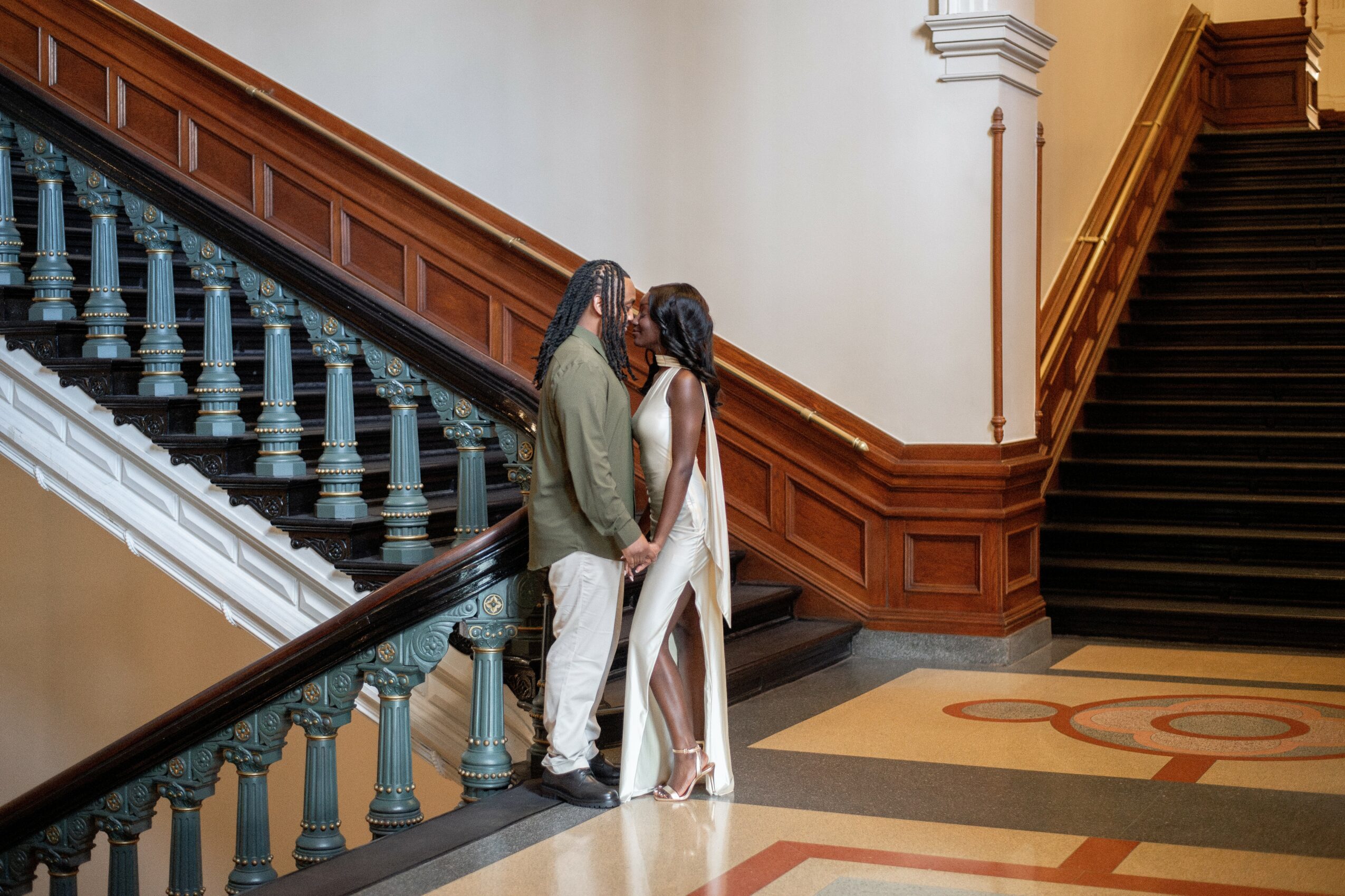A couple stands on a staircase landing, facing each other, in an elegant interior with wooden paneling and a patterned floor.