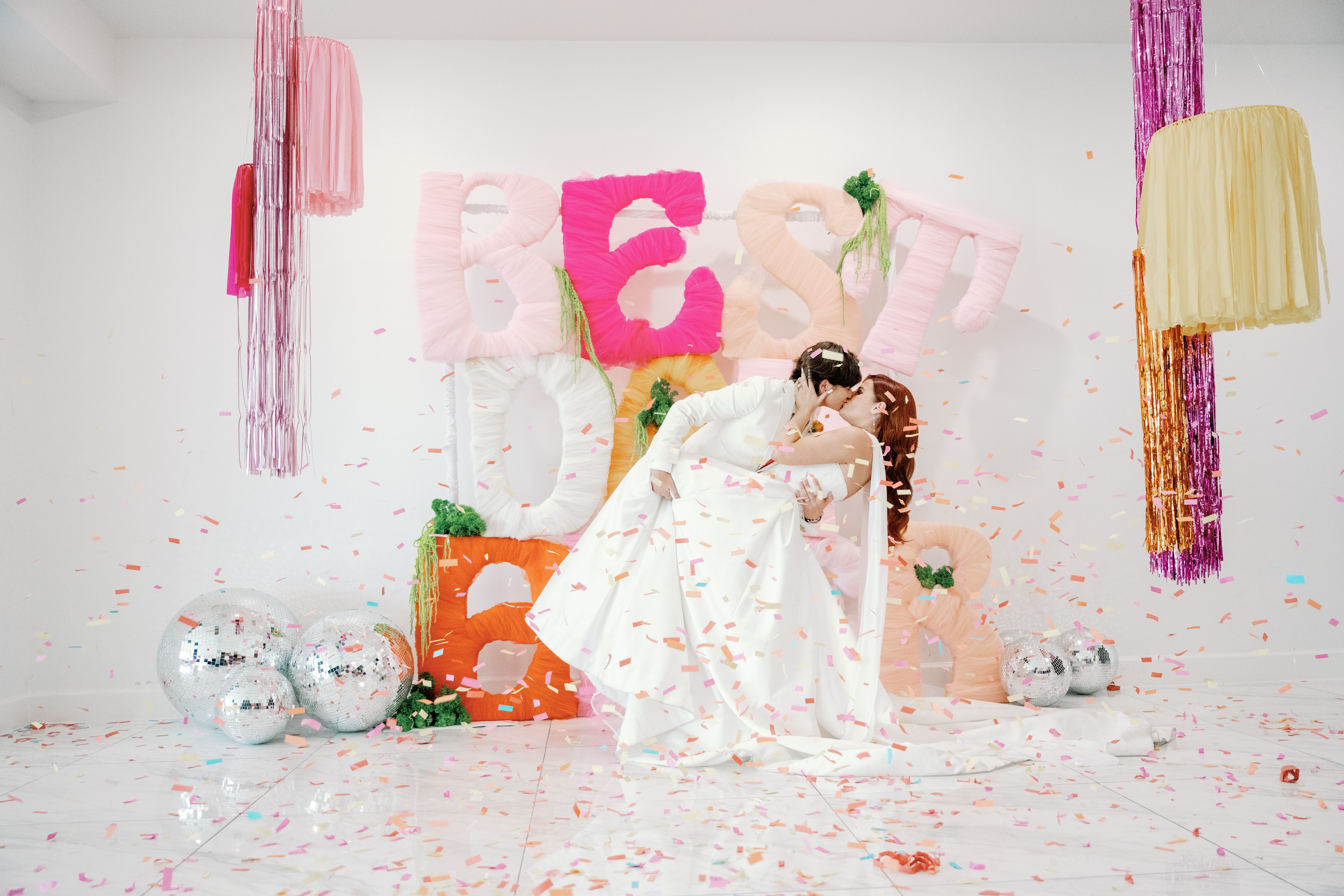Person in a white dress sitting on a pink chair with a decorated backdrop, colorful balloons, and hanging decorations, celebrating a birthday.