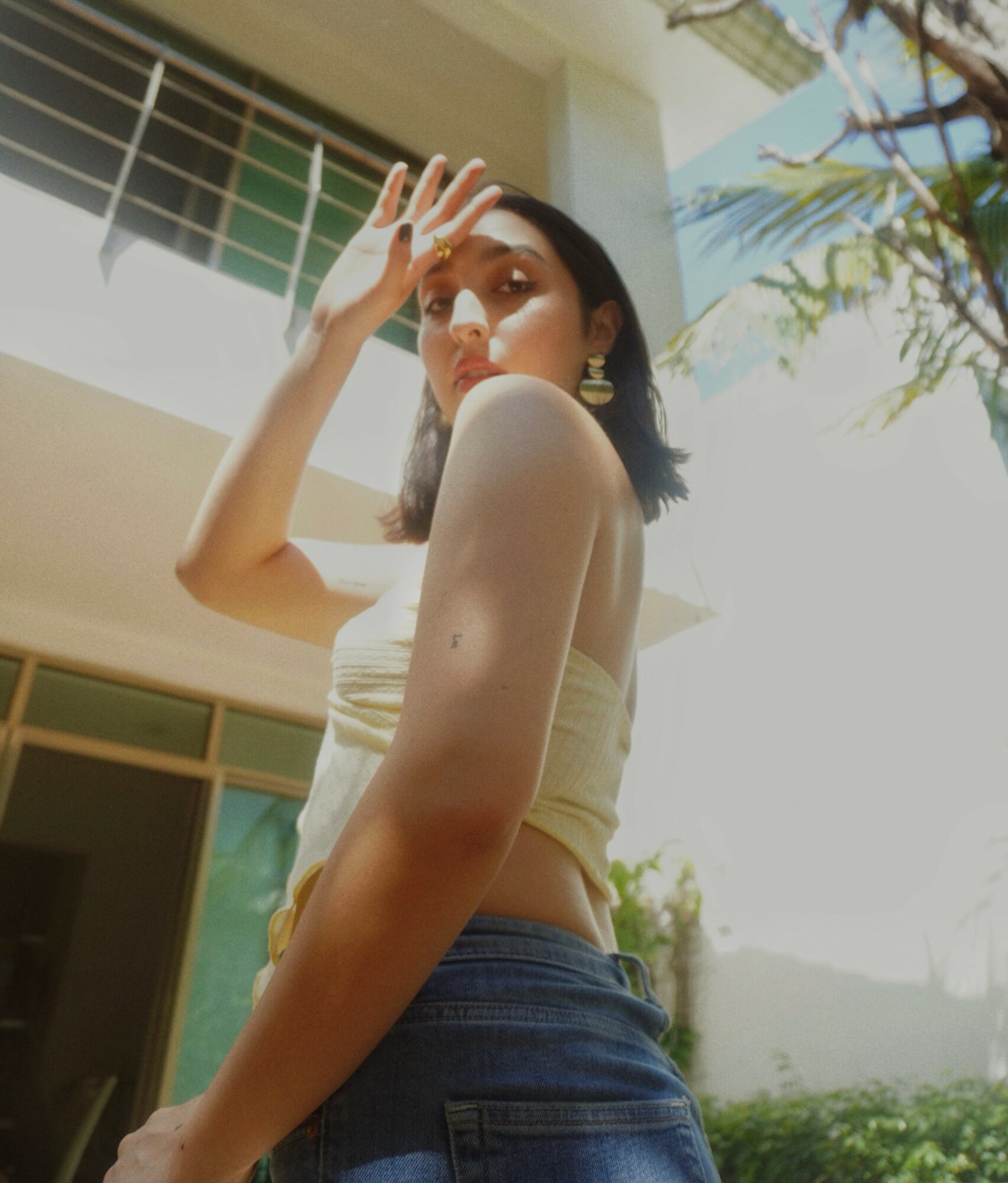 Young woman with short dark hair in a strapless top and jeans outdoors, shielding her eyes from sunlight.