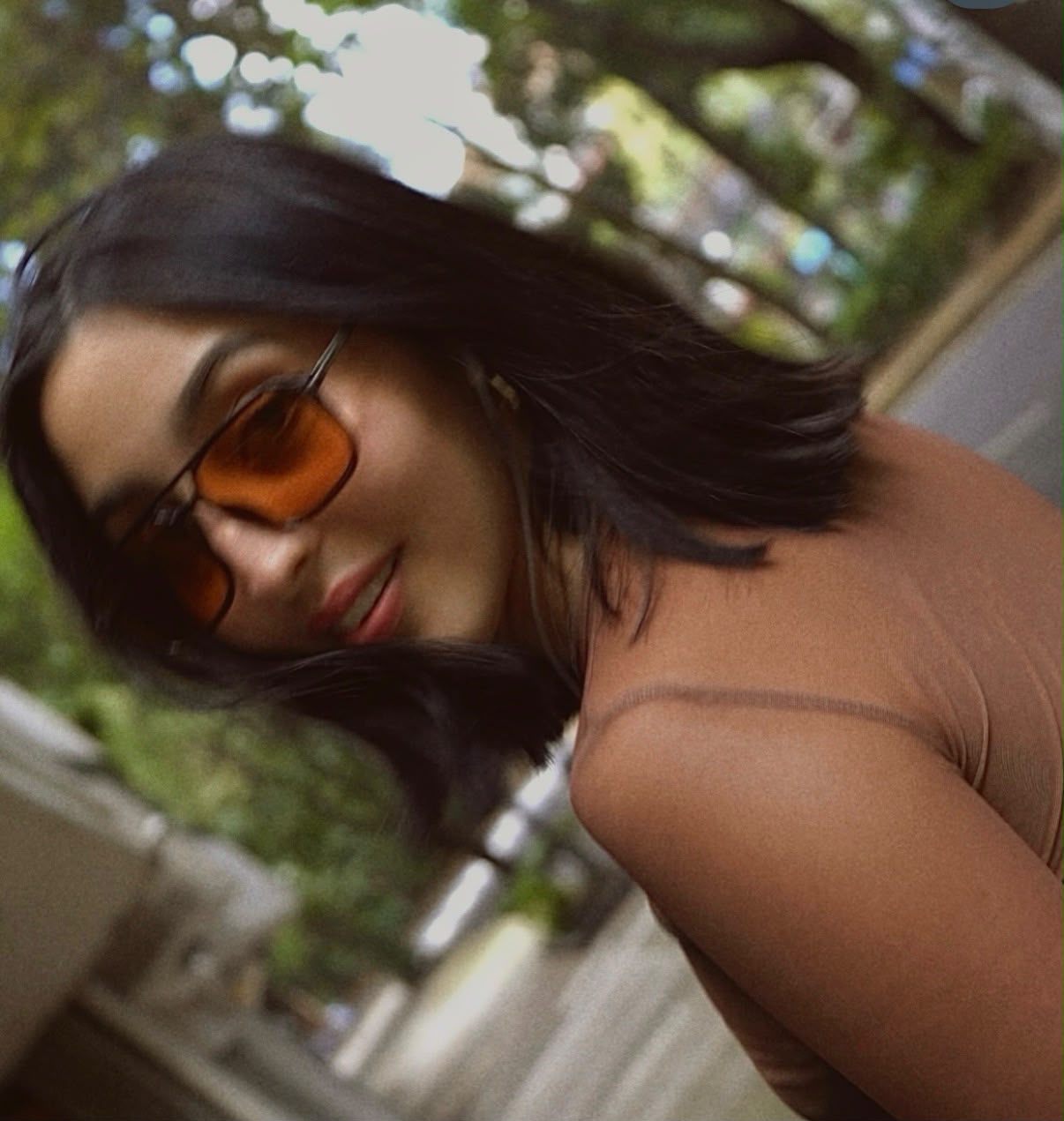 Woman with dark hair wearing sunglasses and a brown top outdoors, smiling slightly, with trees and a building in background.