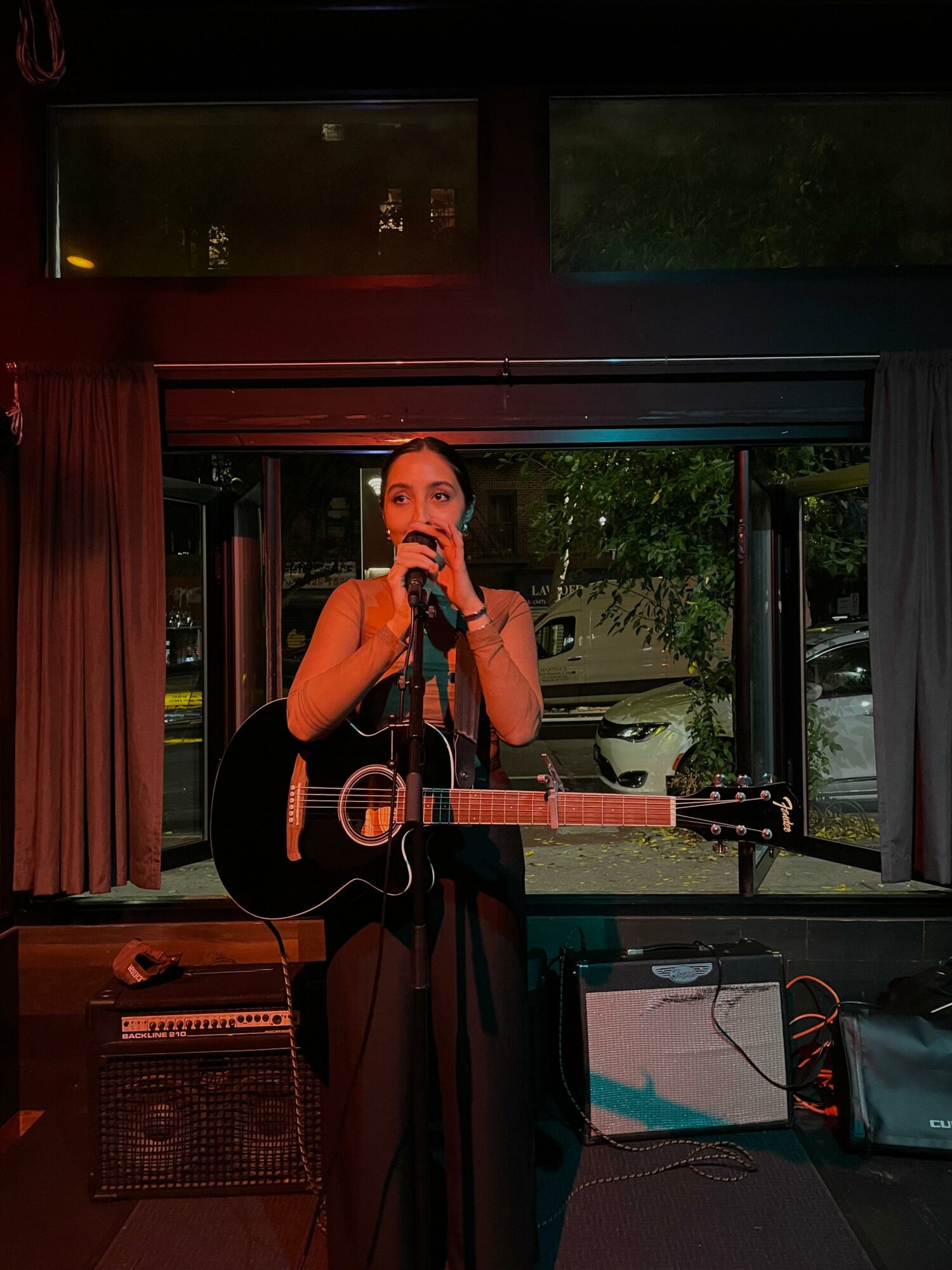 Woman singing into microphone with guitar on stage, indoor setting, equipment visible, curtains and window behind her.
