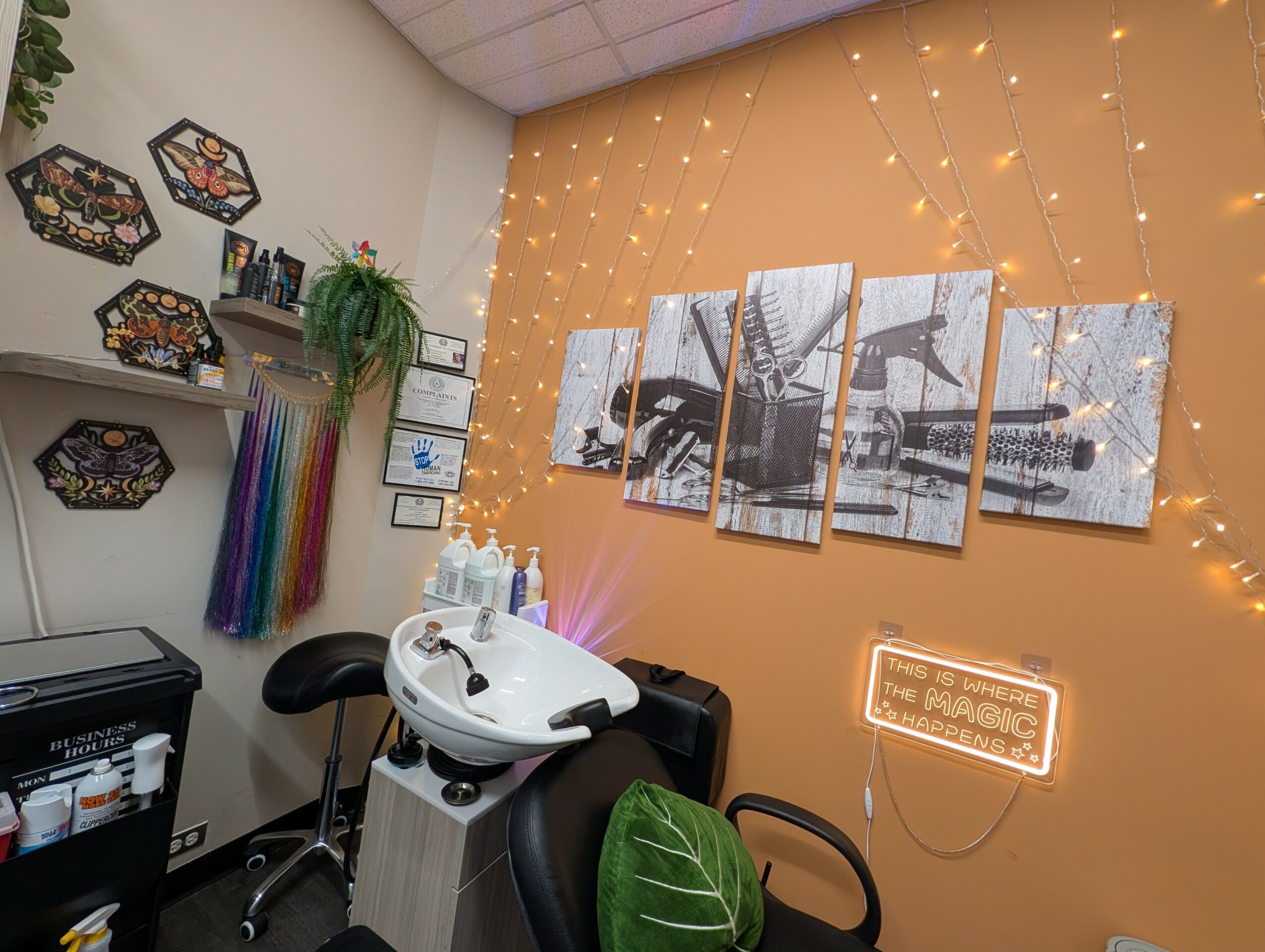 Salon with a hair washing station, decorative wall with fairy lights, framed pictures, and a neon sign, with a chair and plant.