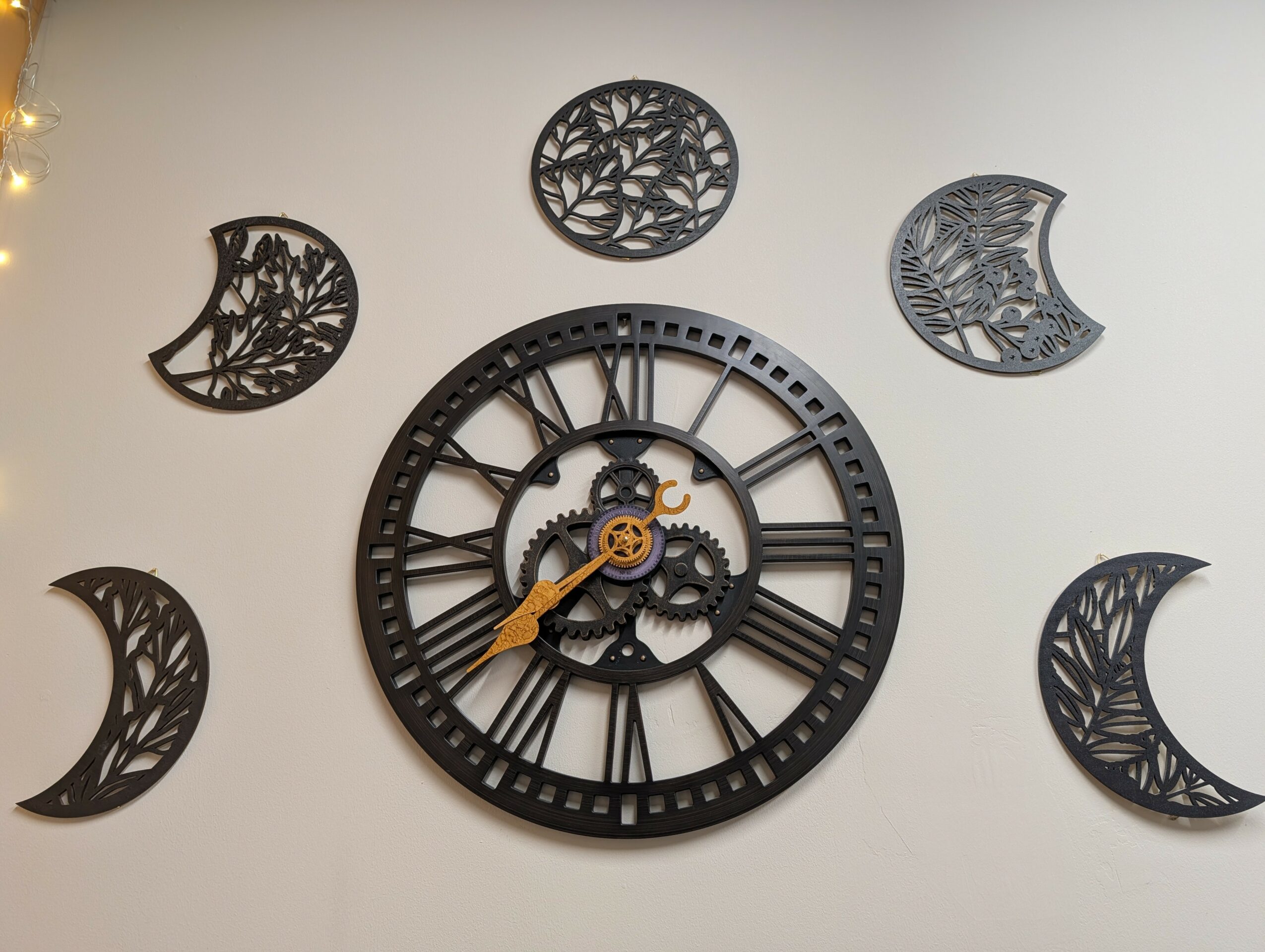 Wall clock with Roman numerals and gear design, surrounded by six decorative moon phases with leaf patterns, on a white wall.