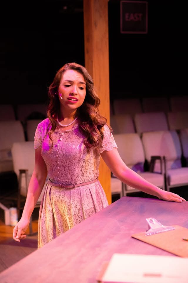 Young woman with long wavy hair in a pink and yellow dress standing at a table in a theater setting.