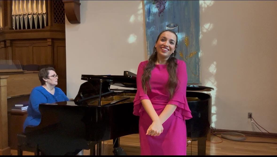 A woman in a pink dress stands smiling in front of a piano, with a person playing piano nearby.