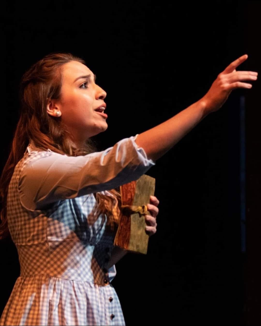 Woman with long hair holding a book, reaching out with her right hand, on a dark stage background.
