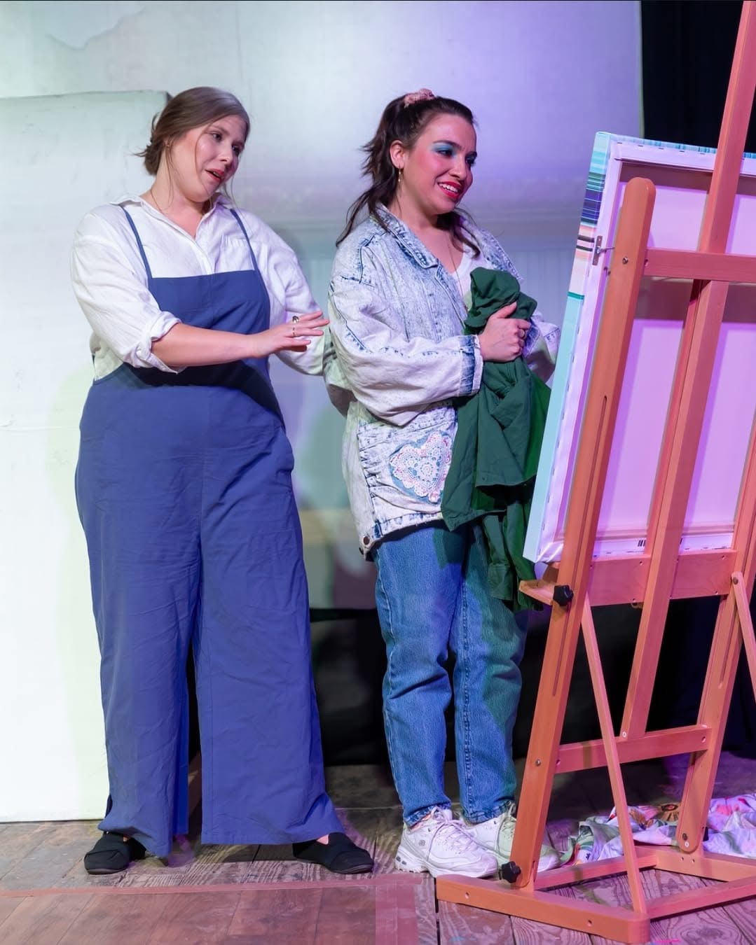 Two women stand near an easel, one smiling and holding a green cloth, the other looking at her, in a room with a plain background.