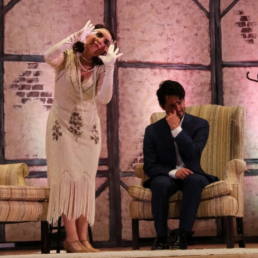 Two people on stage, one standing and gesturing, the other sitting with hand on face, in front of a pink wall.