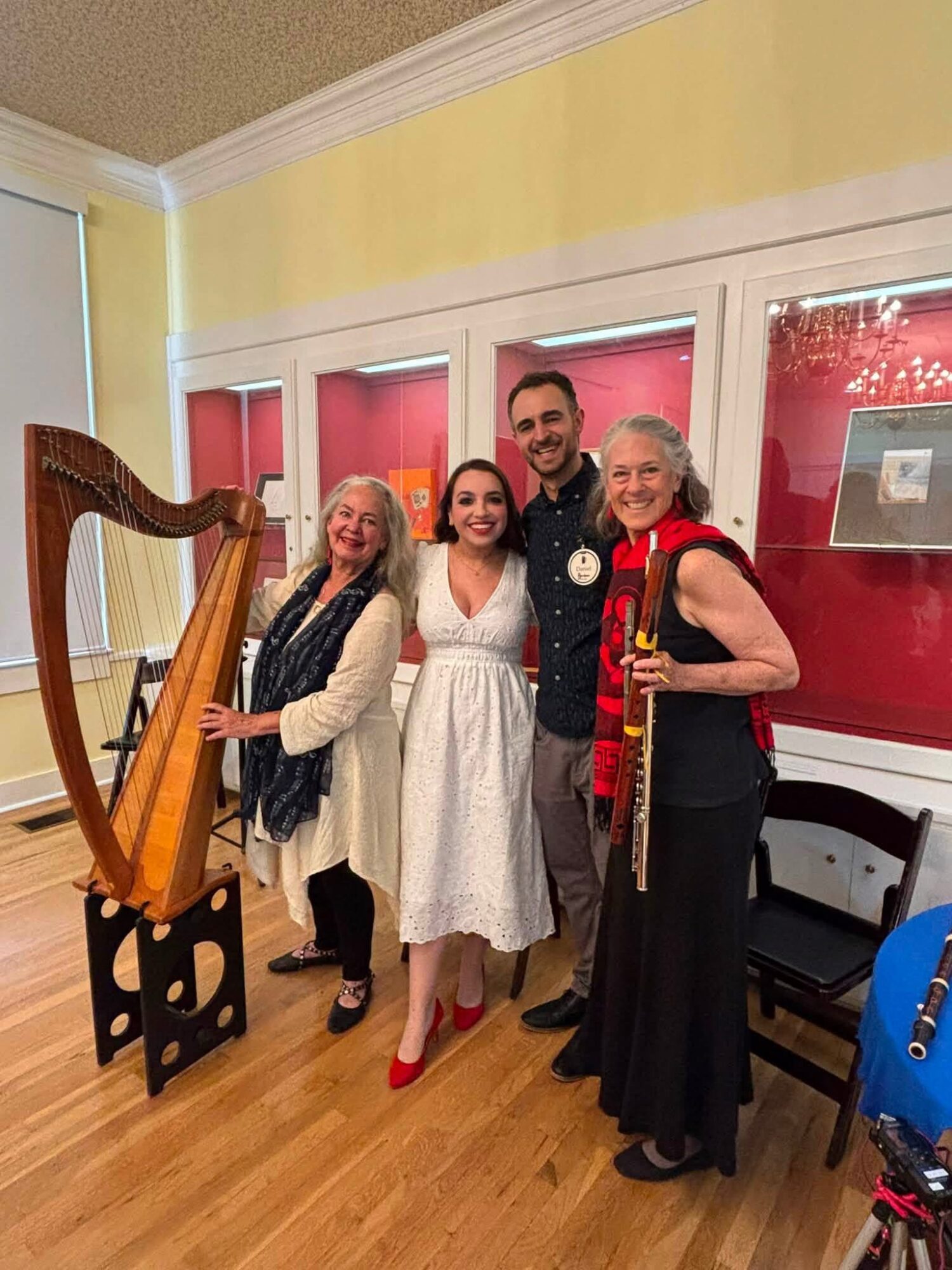 Four people standing indoors, smiling, with a harp on the left and red window blinds behind them.