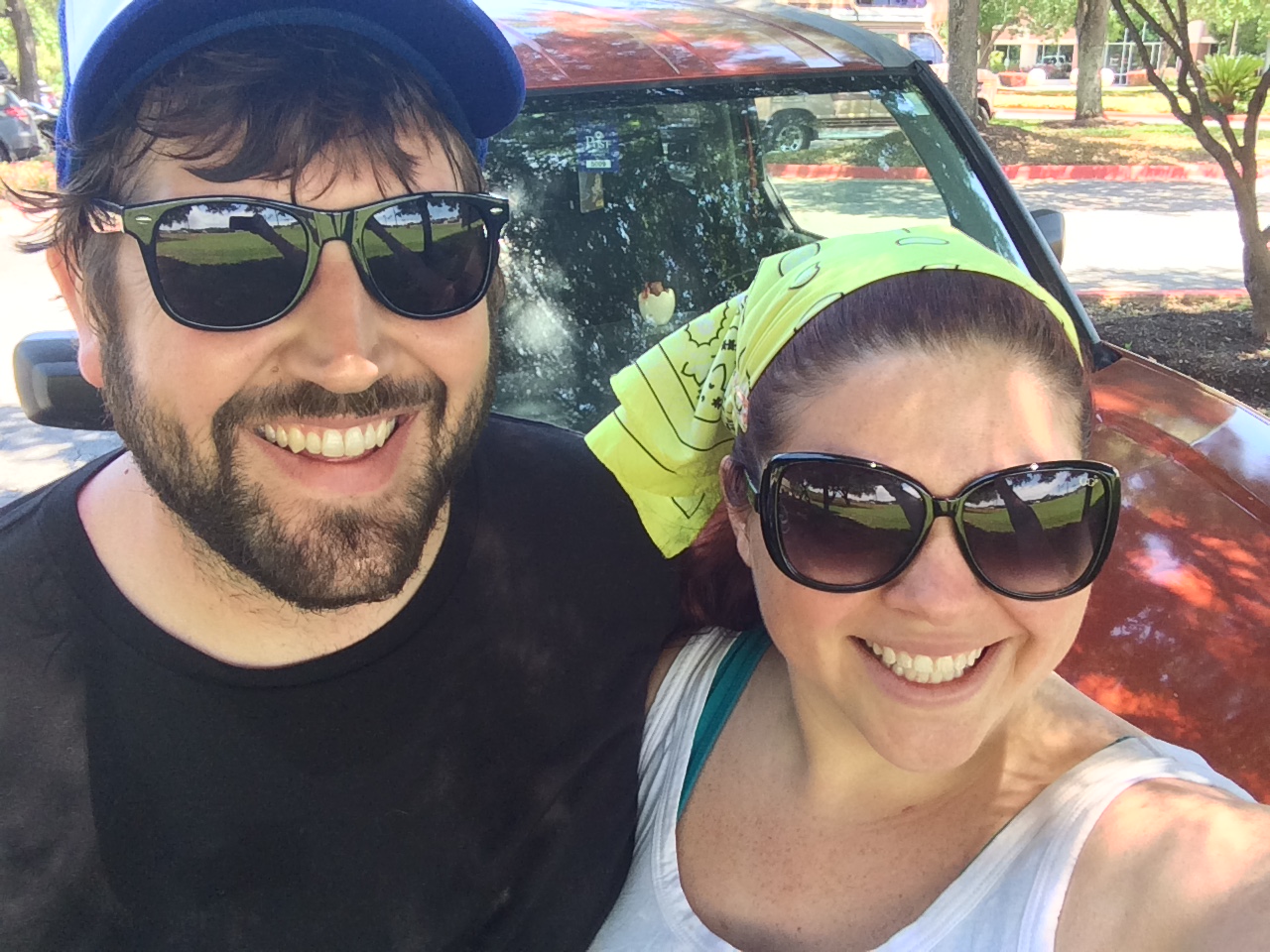 A smiling man and woman wearing sunglasses outdoors, with a car and trees in the background.