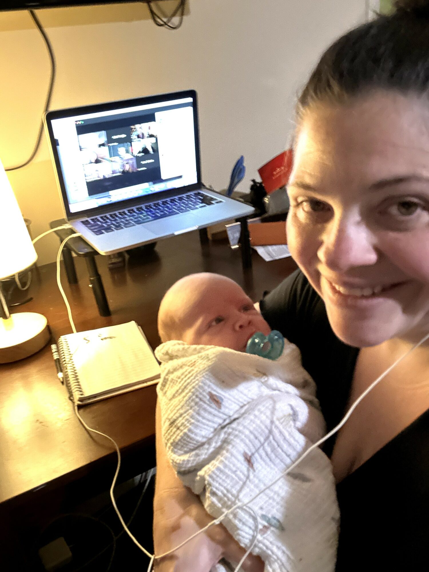 Woman holding a baby with a pacifier, smiling, in a room with a laptop and notebook on a desk.