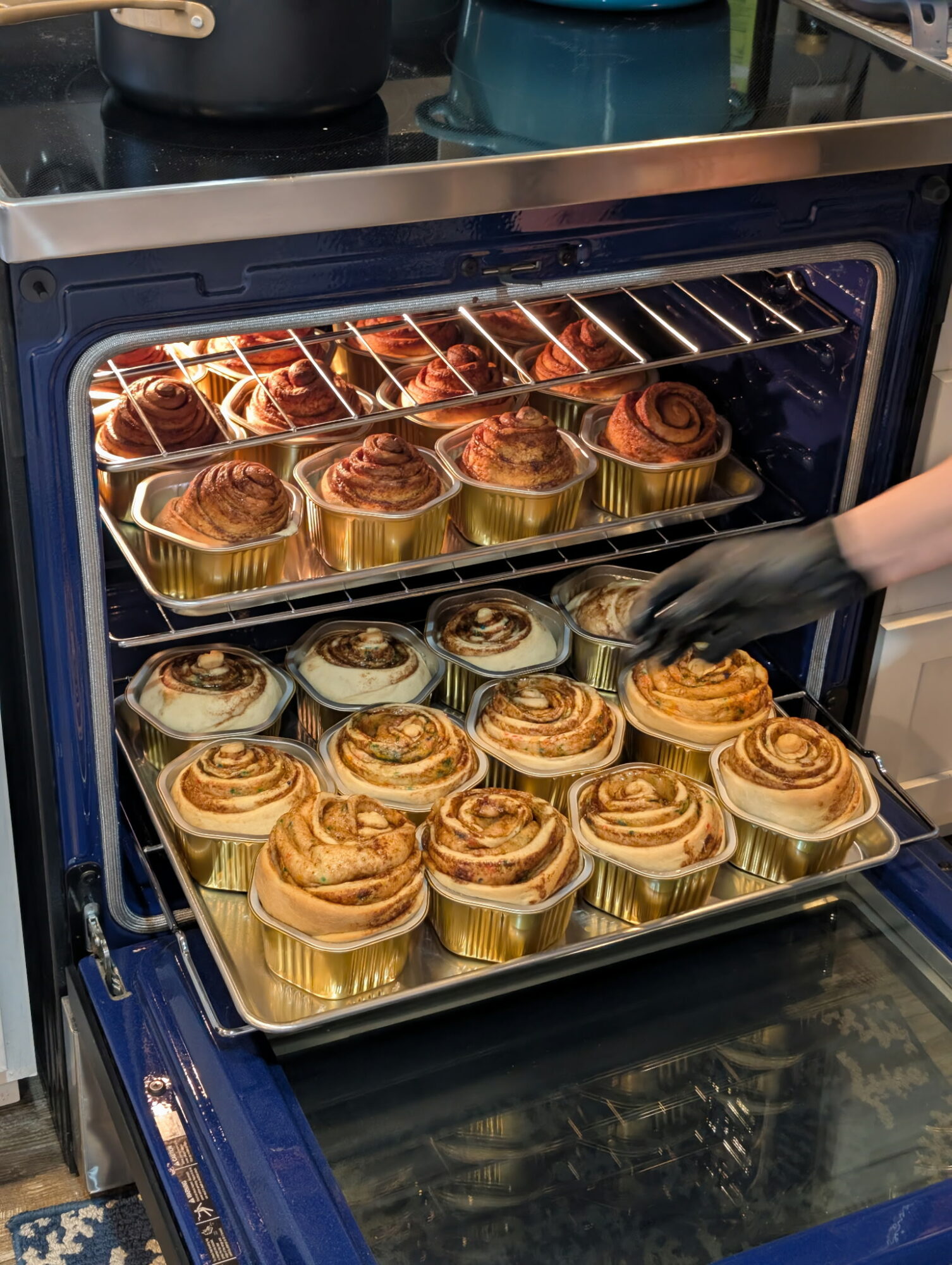 Oven with trays of cinnamon rolls baking inside, some being removed with a gloved hand.