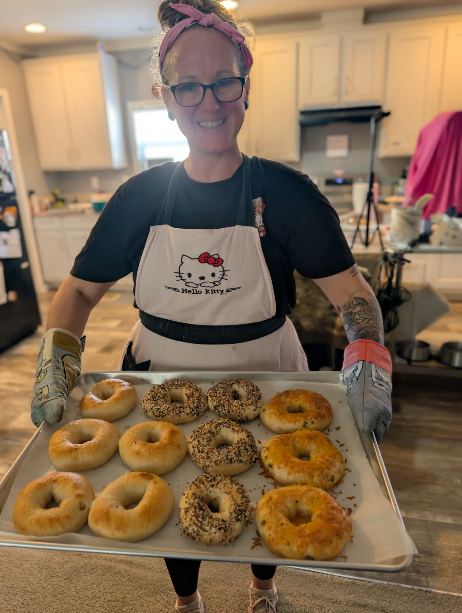 Woman wearing glasses, apron, and gloves holding a tray of assorted bagels in a kitchen.