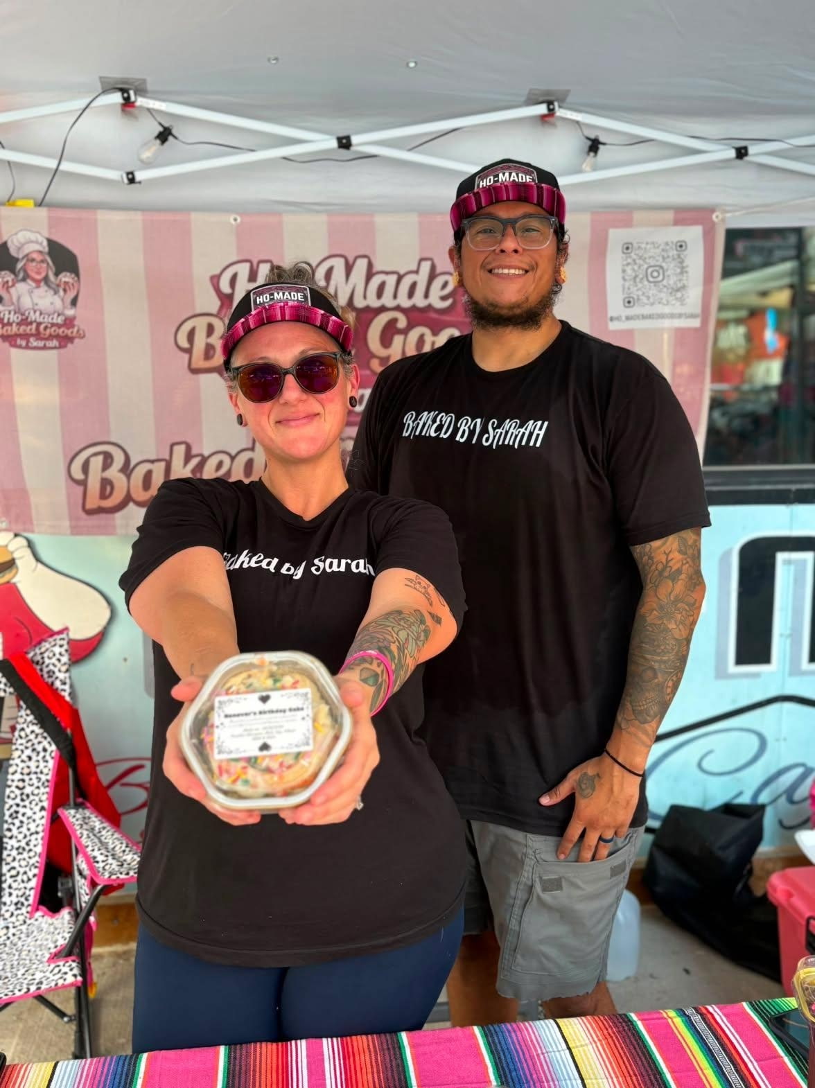 Two people smiling, one holding a cake, standing in front of a pink and white bakery banner, under a canopy.