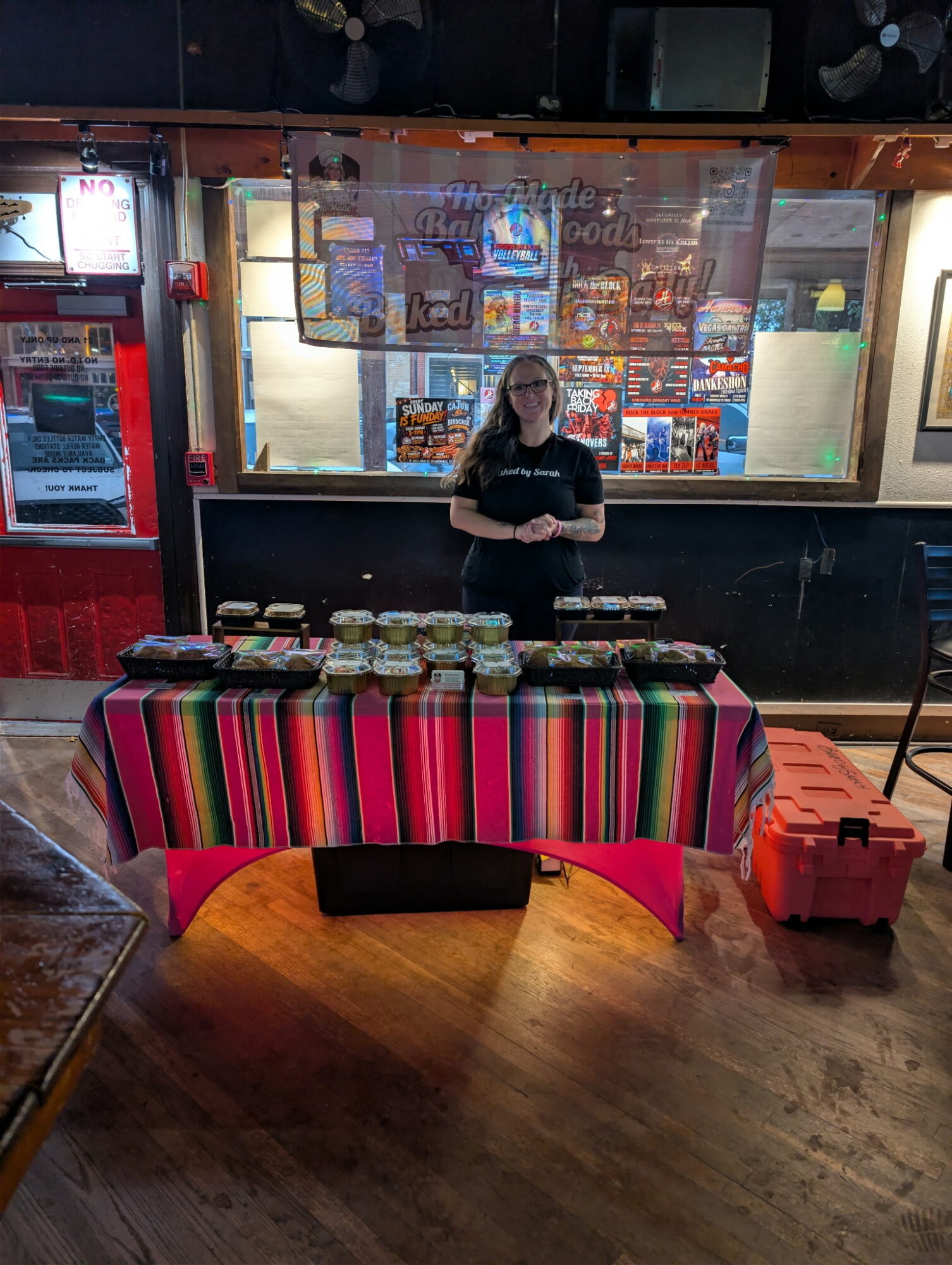 Person standing behind a table with food jars, in front of a window with colorful posters, on a wooden floor.
