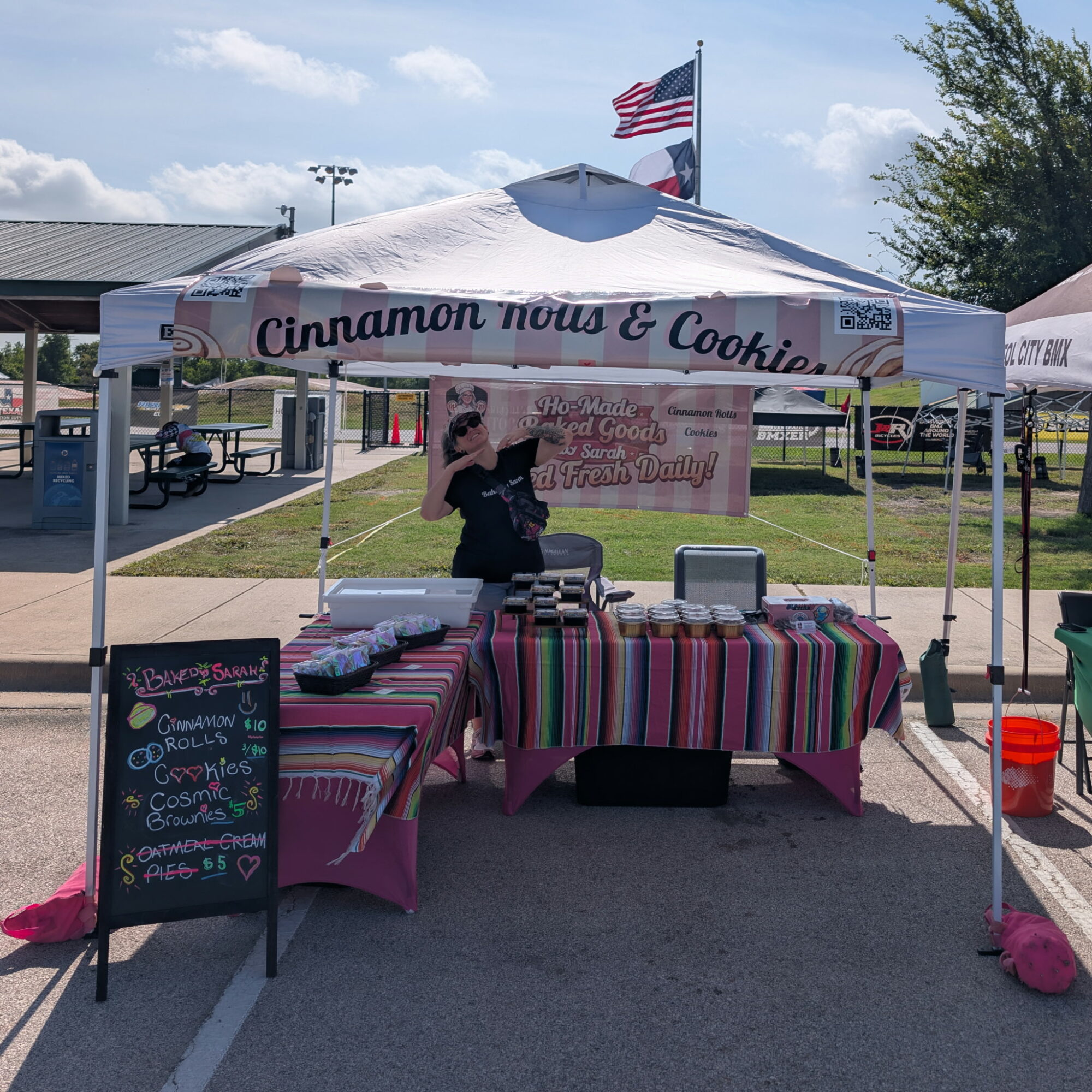 Person standing behind a table under a white tent with a sign reading 'Cinnamon rolls & Cookies' at an outdoor event.