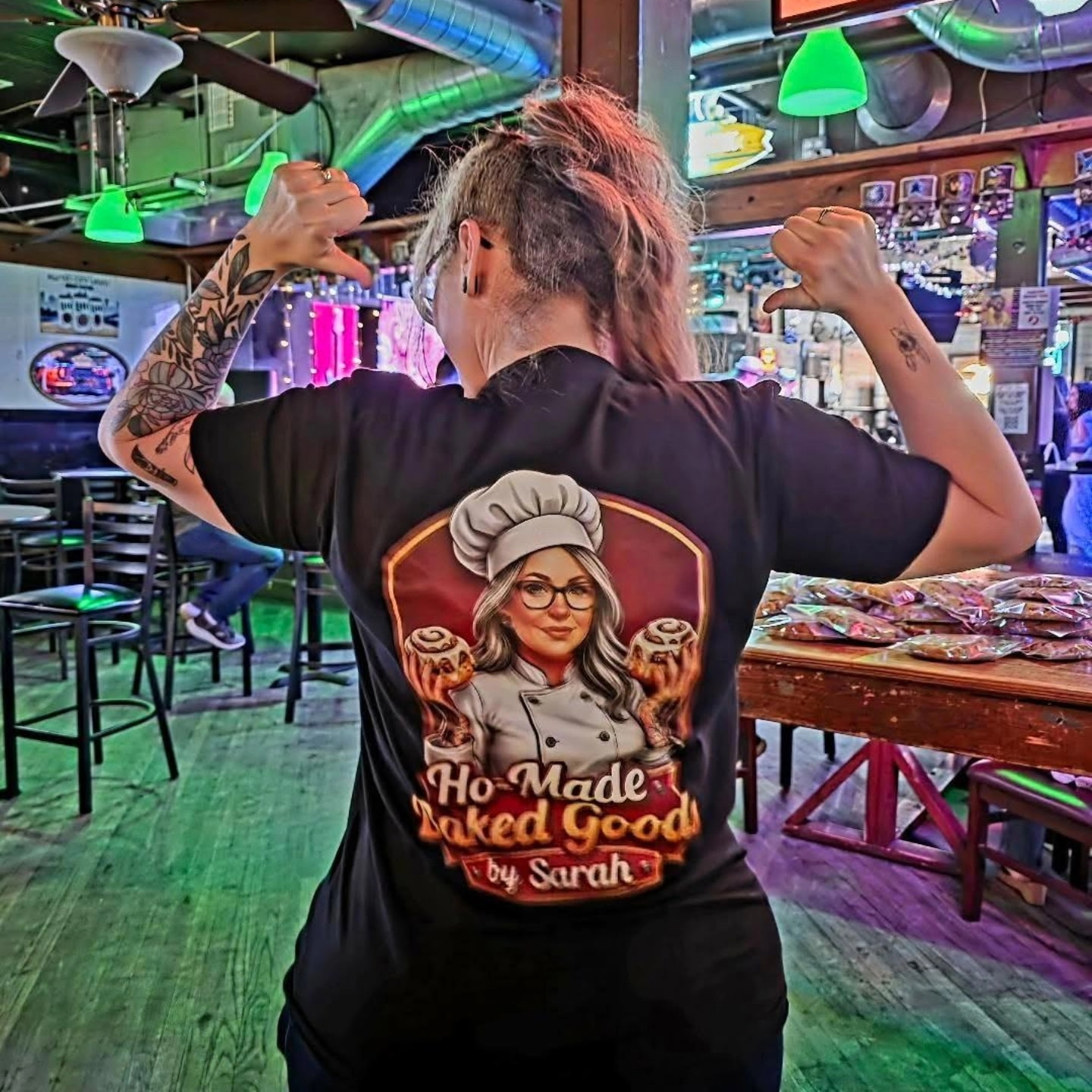 Person with tattooed arms wearing a black T-shirt with a chef illustration and text, in a colorful bar or restaurant.