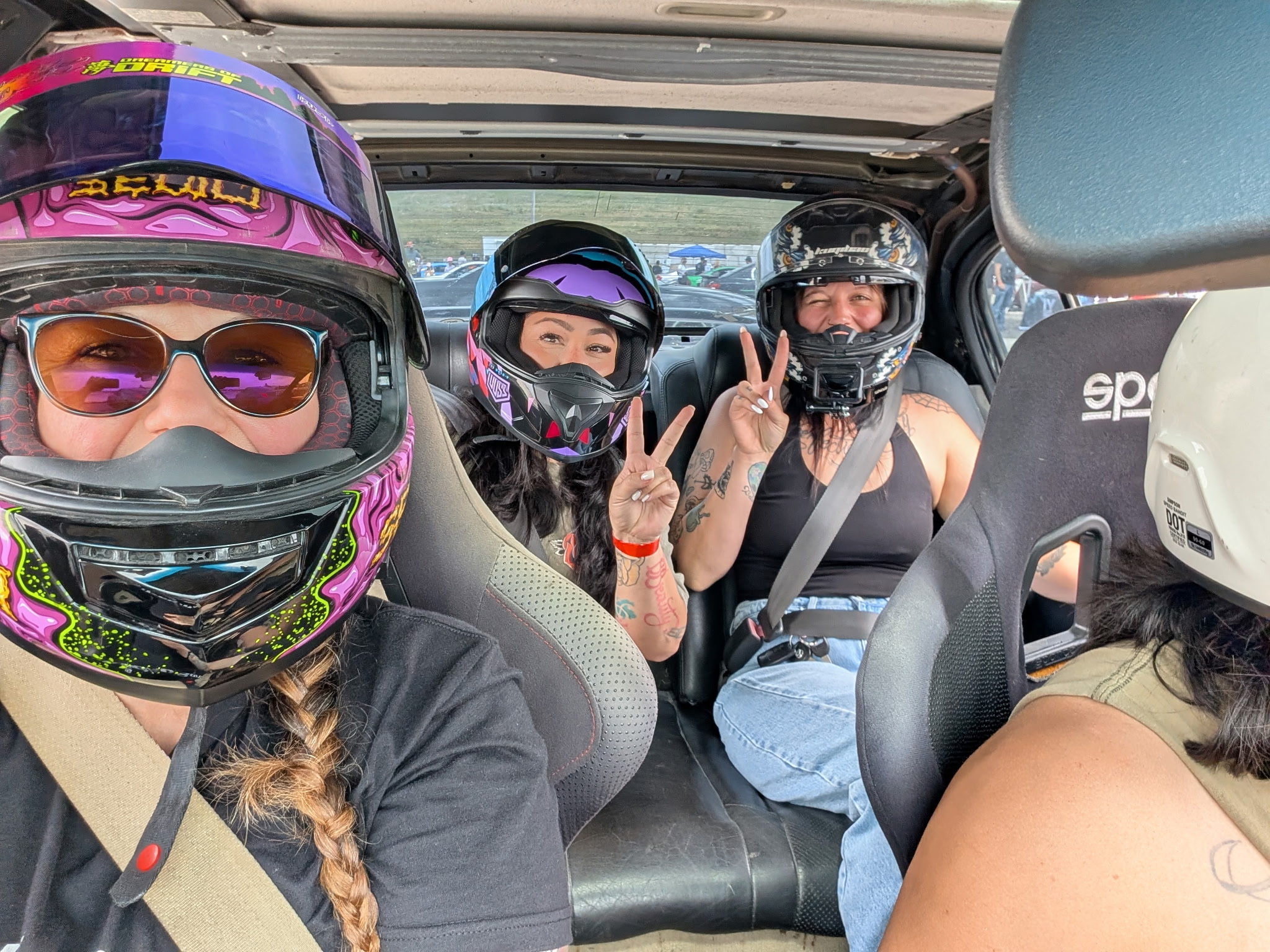 Three people wearing helmets and goggles sitting inside a vehicle, two making peace signs, one smiling.