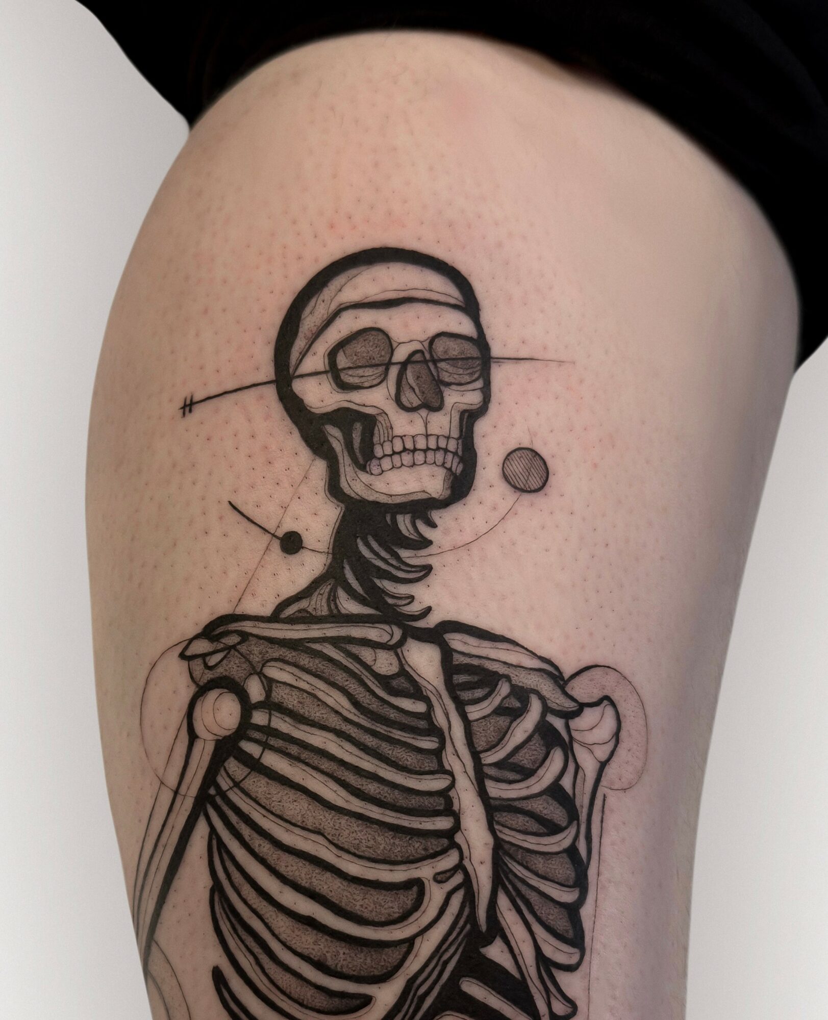 Tattoo of a skull with a line through it, connected to a ribcage, on person's thigh, black ink, detailed line art.
