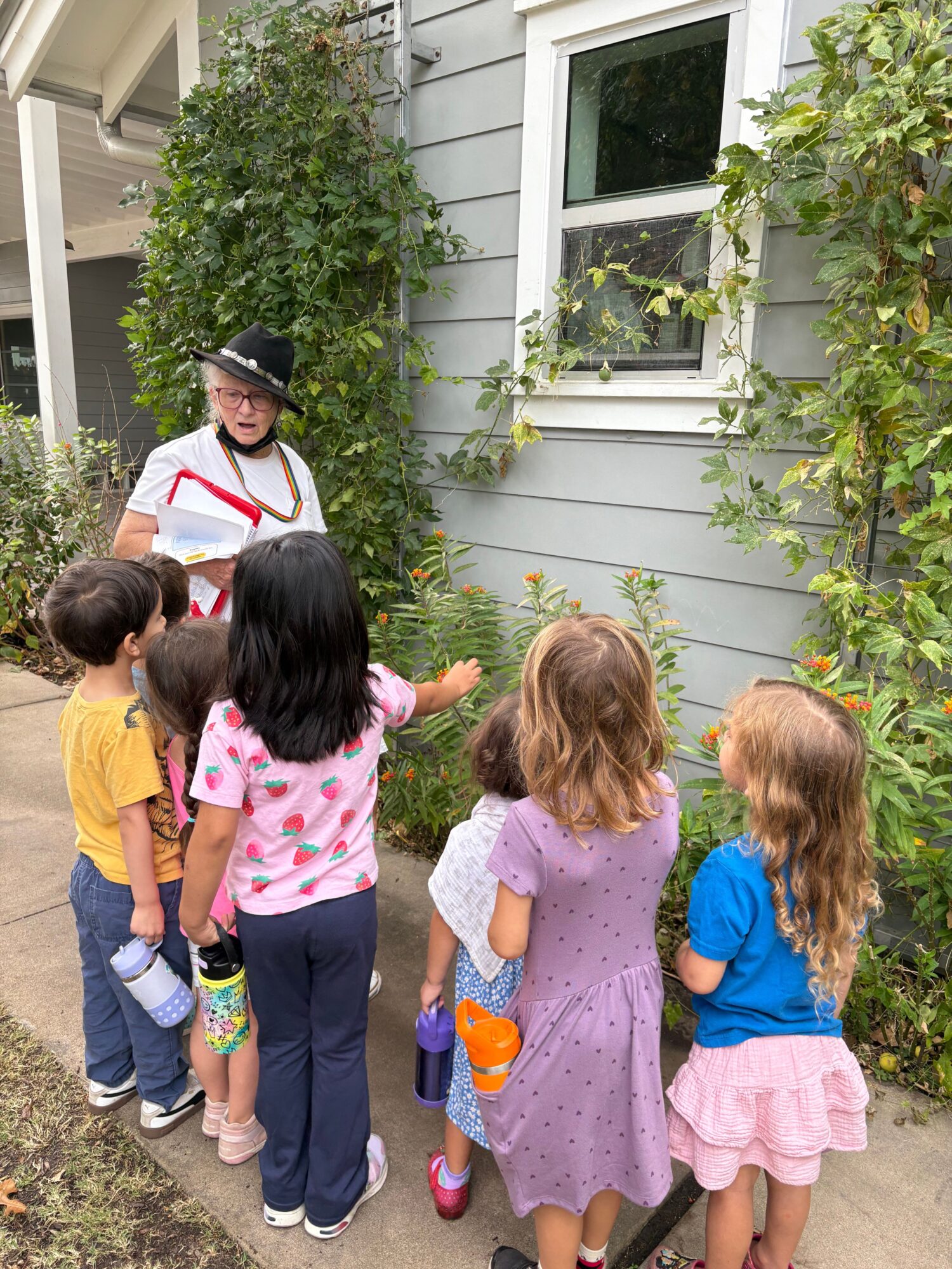 Several children stand beside a house, looking toward a garden path beneath leafy branches.