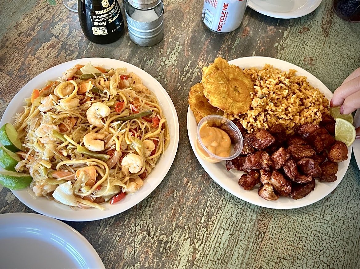 Two plates of food on a wooden table, one with noodles and vegetables, the other with fried chicken, ribs, and lime wedges.