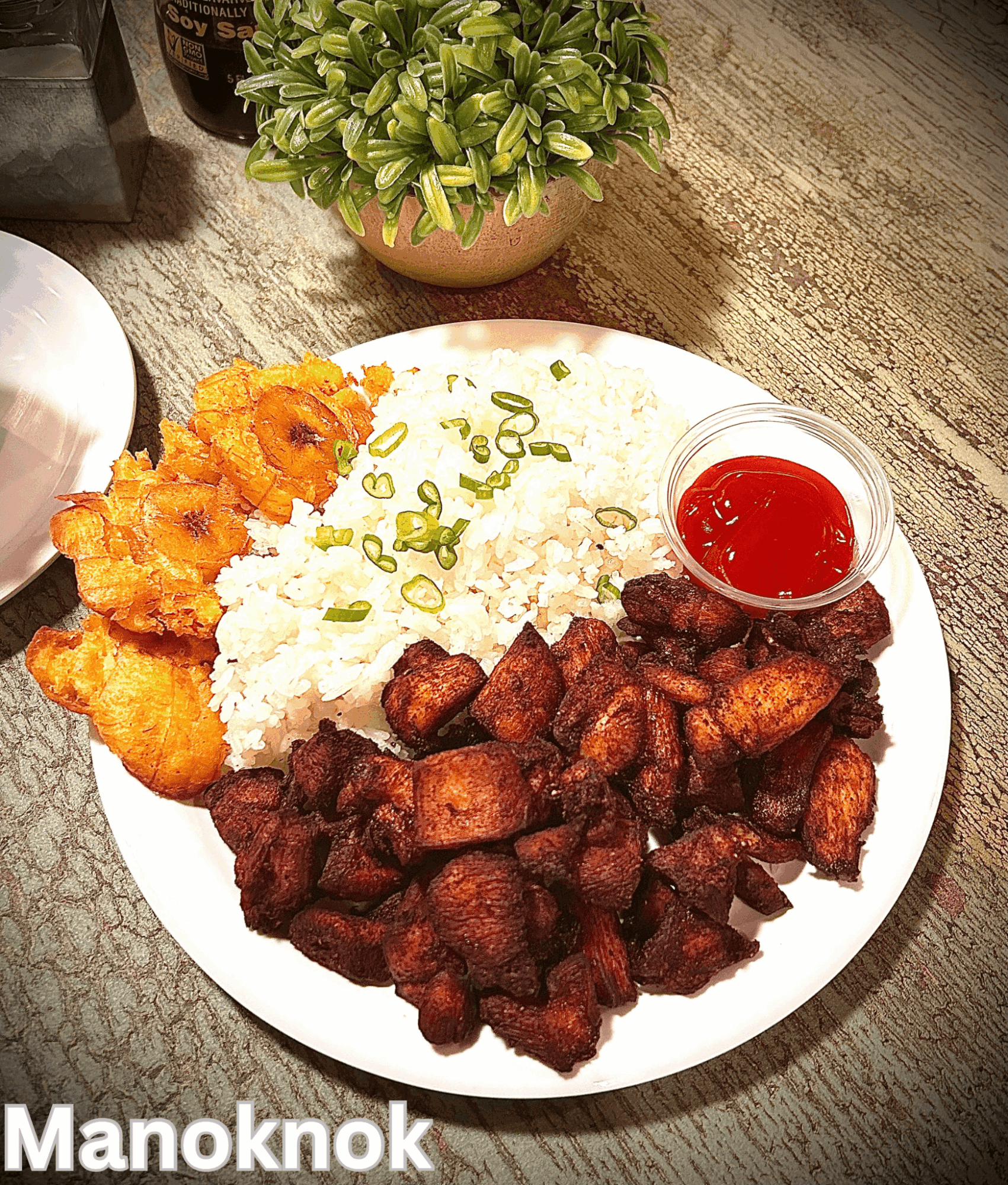 Plate with fried chicken, rice topped with chopped green onions, fried pork, and a small container of red sauce on a wooden table.