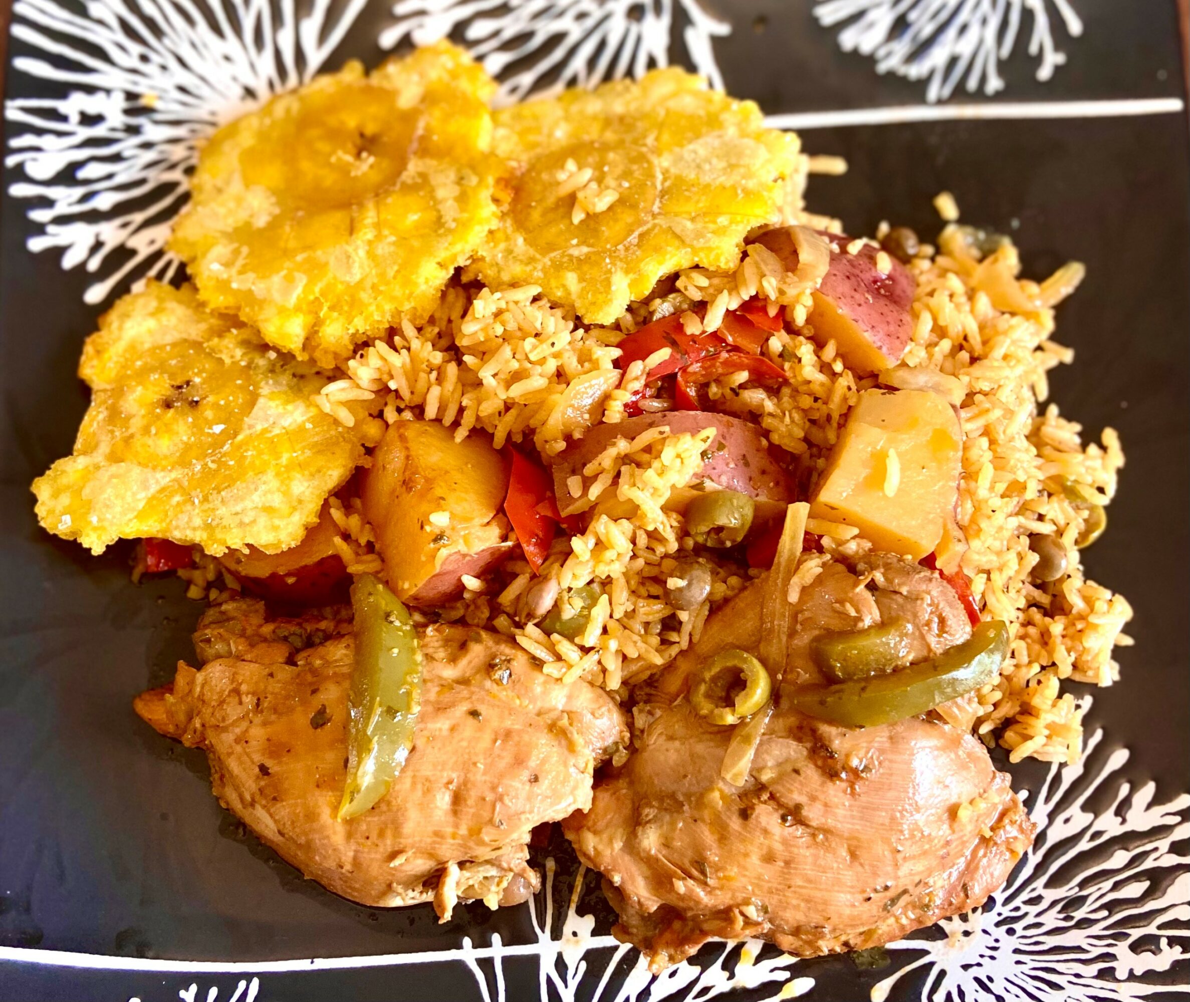 Fried chicken with rice, vegetables, and fried plantains on a black plate with white decorative patterns.