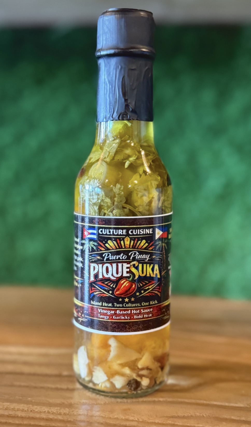 Bottle of Pique Suka sauce with yellowish contents and a black cap, on a wooden surface with green background.