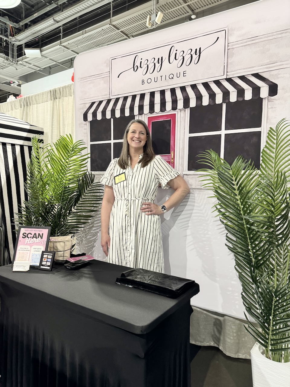 Woman standing in front of a boutique backdrop with plants on sides, smiling, wearing a striped dress, with a table in front.