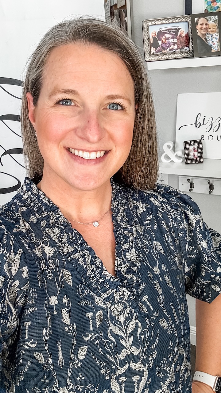 Smiling woman with shoulder-length hair wearing a patterned blouse, in a room with framed photos and decor