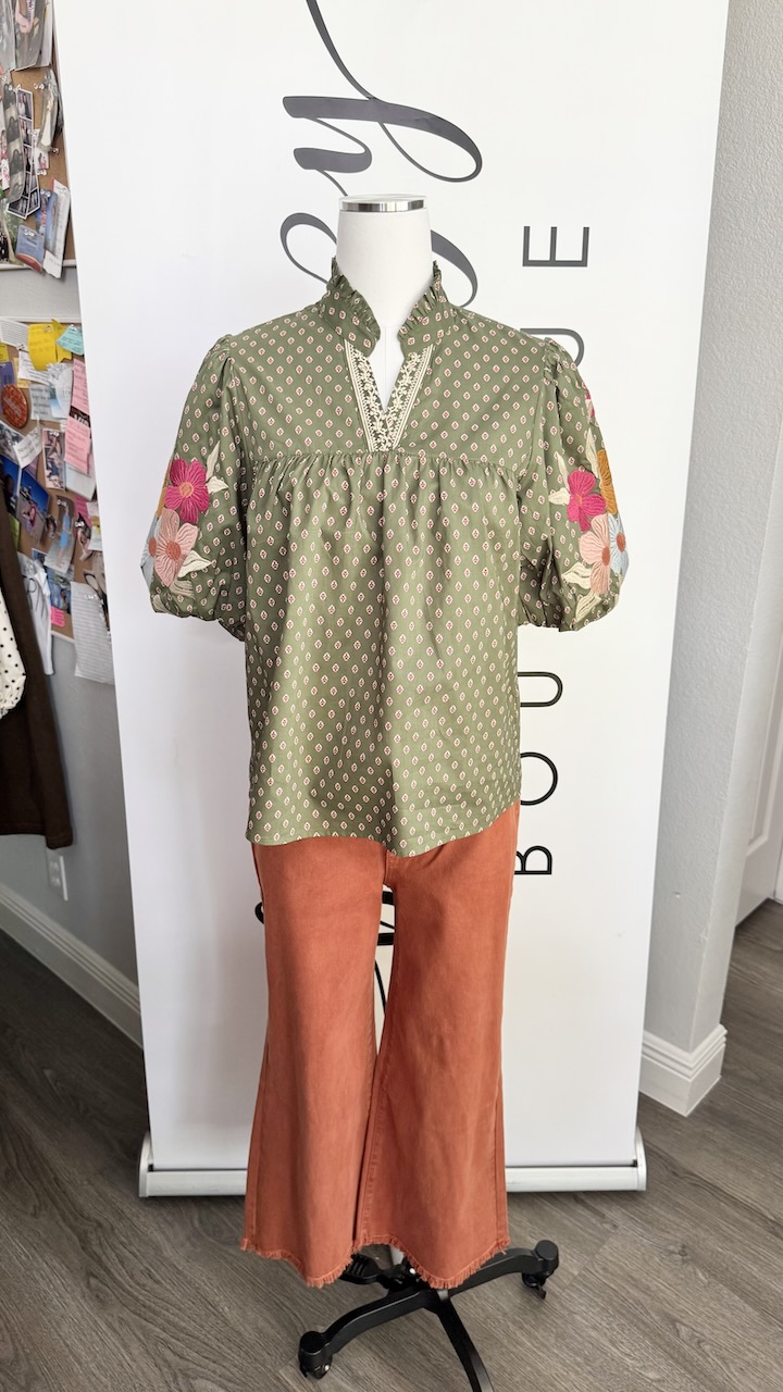 Mannequin wearing a green patterned tunic with pink floral sleeves and orange wide-leg pants, standing in front of a white background.
